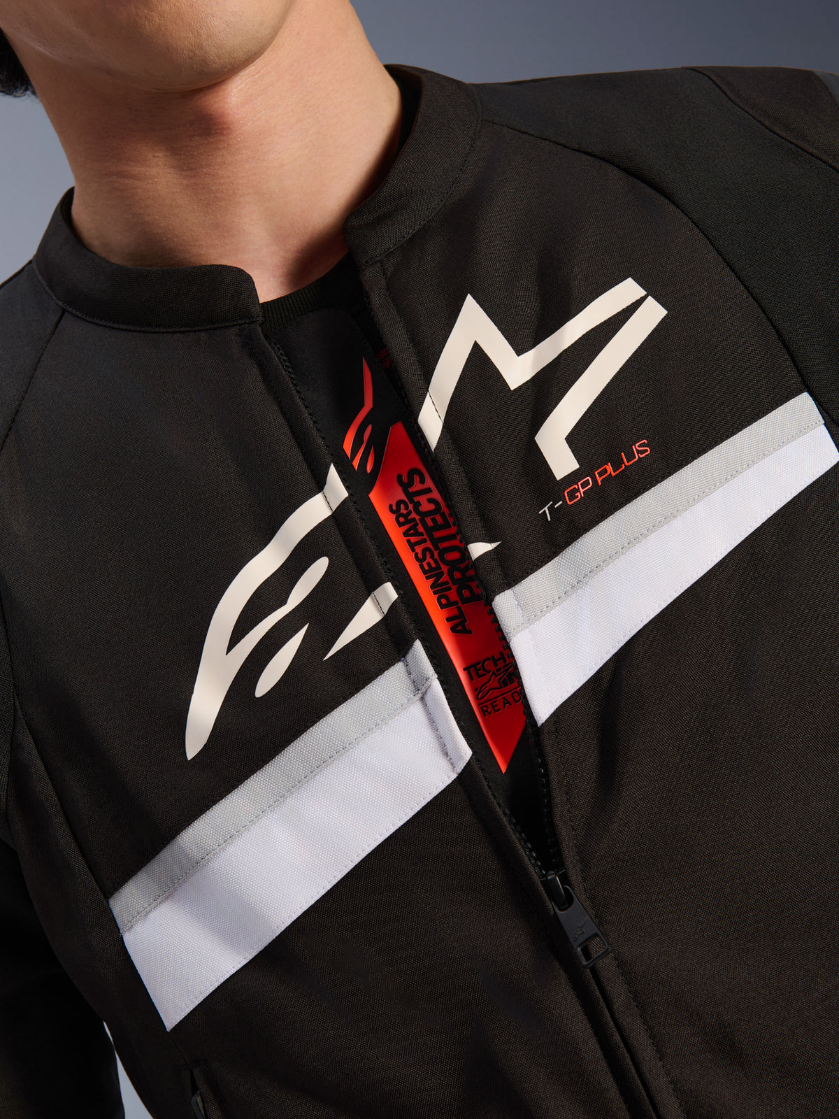 Alpinestars T-GP Plus R V4 Jacke, Nahaufnahme von Brust und Kragen, schwarzes Textil mit weißem Stern-Logo und T-GP PLUS-Branding, weißer und grauer horizontaler Streifen, geöffneter Reißverschluss zeigt rote Innenlasche mit Tech-Air Ready-Schriftzug