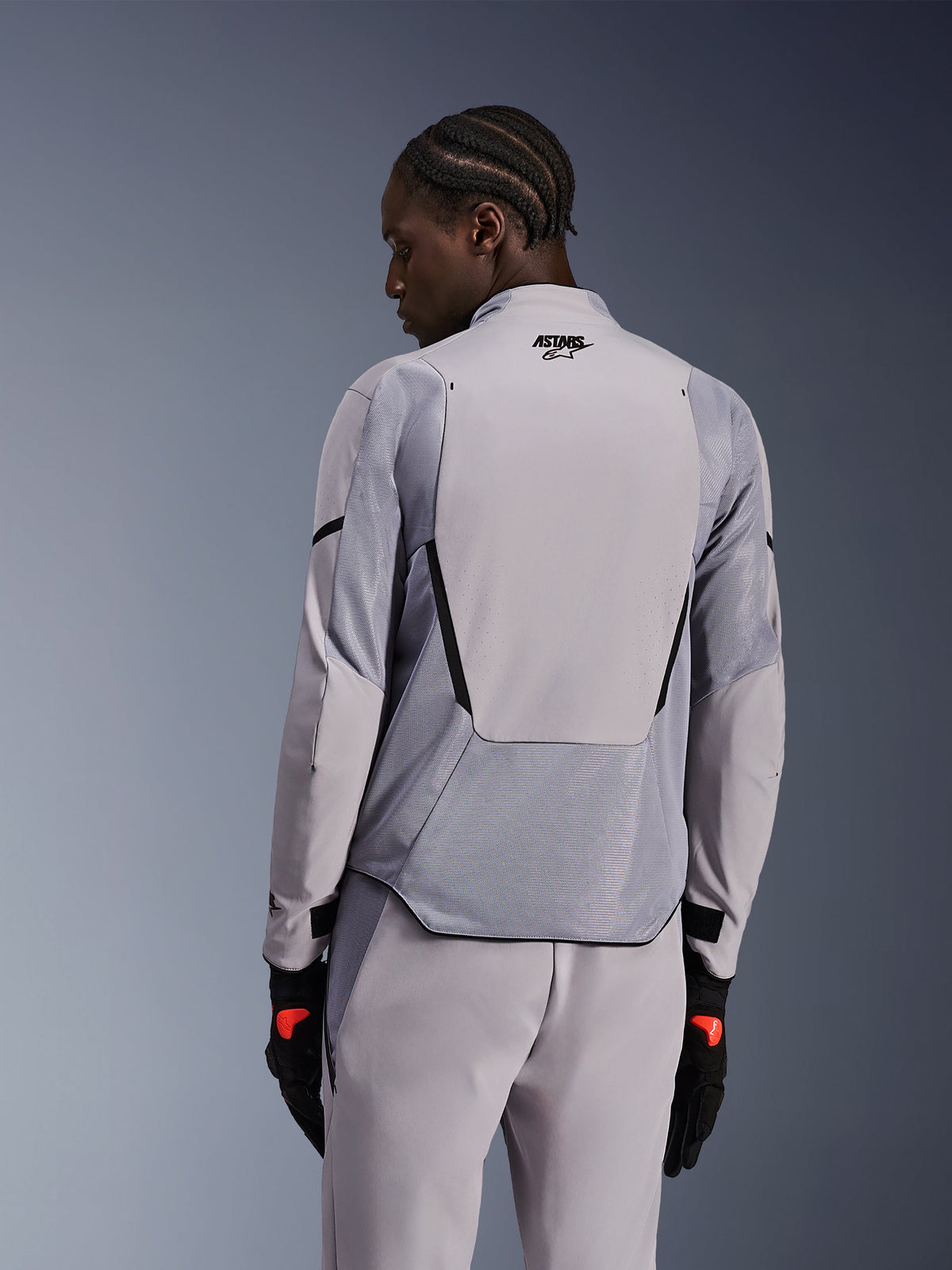 Alpinestars Aeroshell Airflow Jacke in Mid Gray Anthracite, Rückansicht mit atmungsaktiven Mesh-Einsätzen und Laserperforationen, technische Stretch-Softshell-Konstruktion mit verlängertem Rückenteil, kombiniert mit passender grauer Hose und schwarzen Handschuhen für einen sportlichen Look.