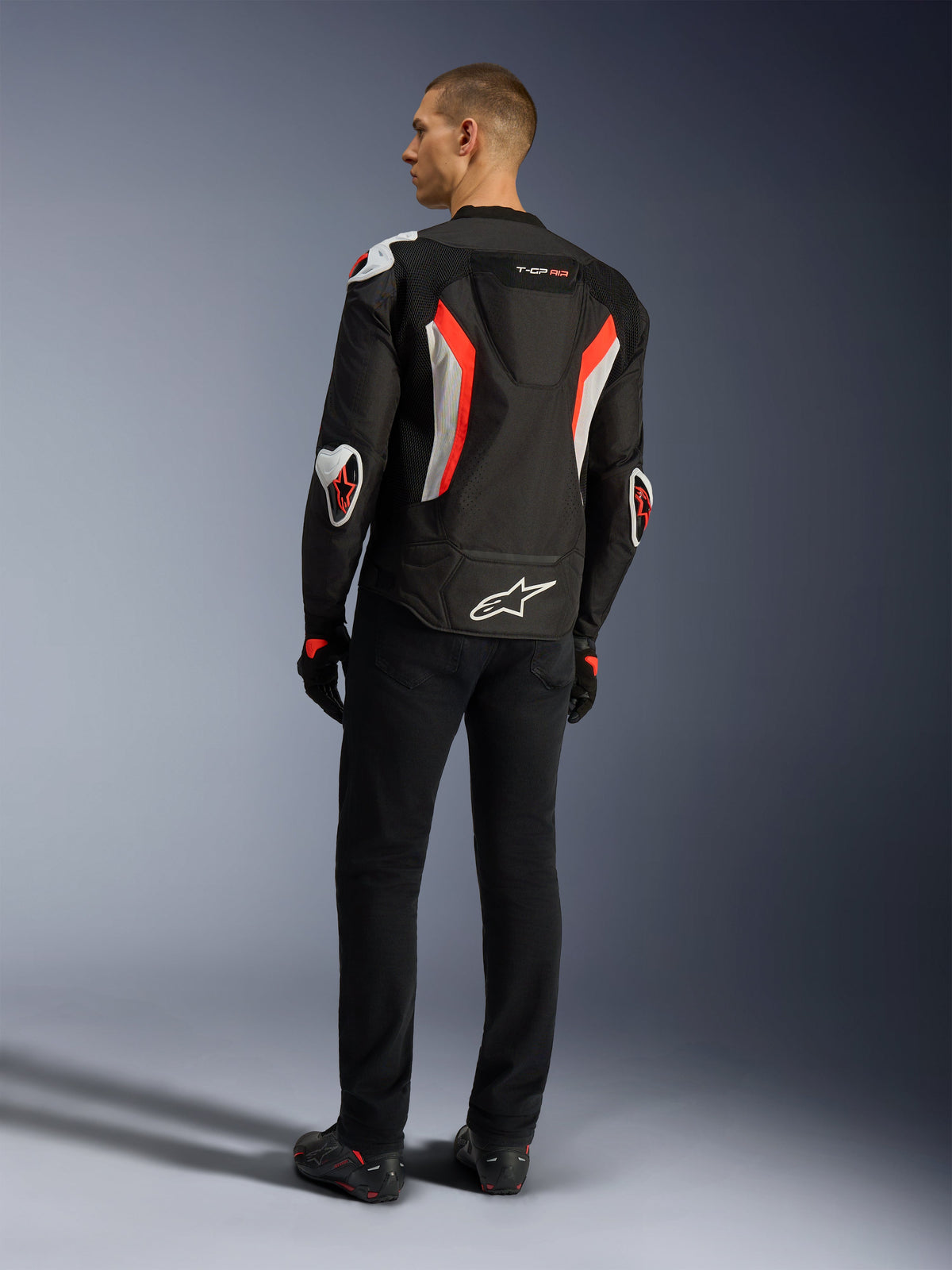 Alpinestars T-GP Air Jacke, Motorradjacke in Schwarz und Fluo-Rot, Rückansicht an einem Model, mit Mesh-Belüftungseinsätzen, aerodynamischem Höcker und weißen externen Protektoren an Ellbogen und Schultern, getragen mit schwarzer Hose und Motorradschuhen.