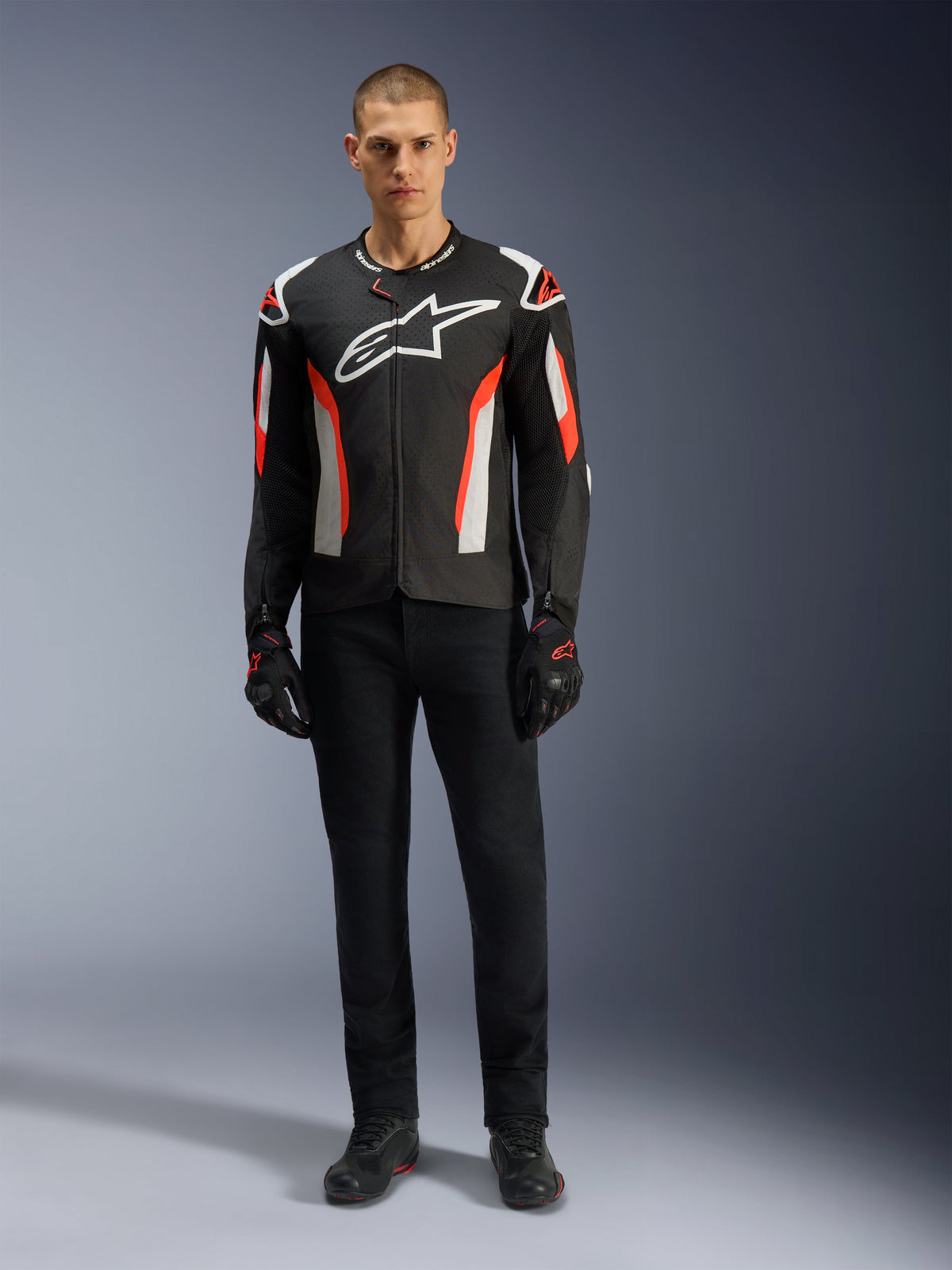 Männliches Model trägt die Alpinestars T-GP Air Jacke in Schwarz-Rot-Fluo, Textil-Motorradjacke mit weißen und leuchtend roten Akzenten, großes weißes Alpinestars-Logo auf der Brust, mit Mesh-Belüftungseinsätzen, kombiniert mit einer schwarzen technischen Hose und Motorradhandschuhen, sportlich geschnittene Ausrüstung