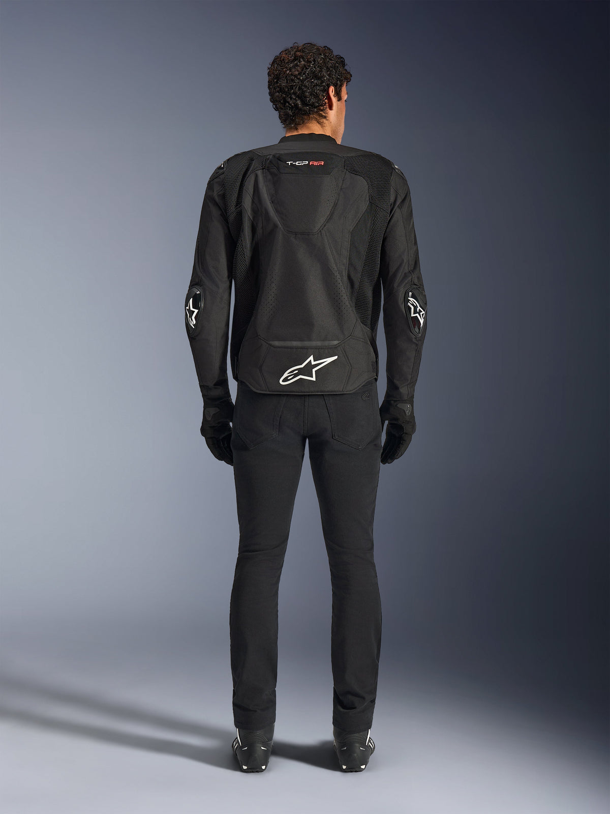 Alpinestars T-GP Air Jacke, Schwarz, Rückansicht eines Mannes, der die Textil-Motorradjacke trägt, mit Mesh-Belüftungseinsätzen, einem aerodynamischen Höcker und weißen Alpinestars-Logos am unteren Rücken und an den Ellbogen, kombiniert mit einer schwarzen Motorradhose und Handschuhen