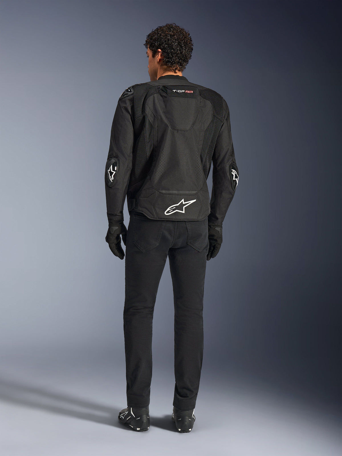 Alpinestars T-GP Air Jacke, Schwarz, Rückansicht am Modell, zeigt 3D-Mesh-Belüftungspanele und aerodynamischen Höcker, mit weißem Stern-Logo am unteren Rücken und an den Ellbogen, getragen mit schwarzer Motorradhose und Handschuhen, sportliche Fahrbekleidung.