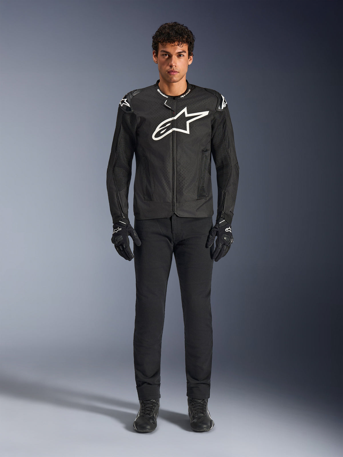 Alpinestars T-GP Air Jacke, schwarze Textil-Motorradjacke mit einem großen weißen Astars-Logo auf der Brust, getragen von einem Modell mit schwarzer Motorradhose und Handschuhen, mit perforierten Paneelen und Mesh-Belüftung für sportliches Fahren