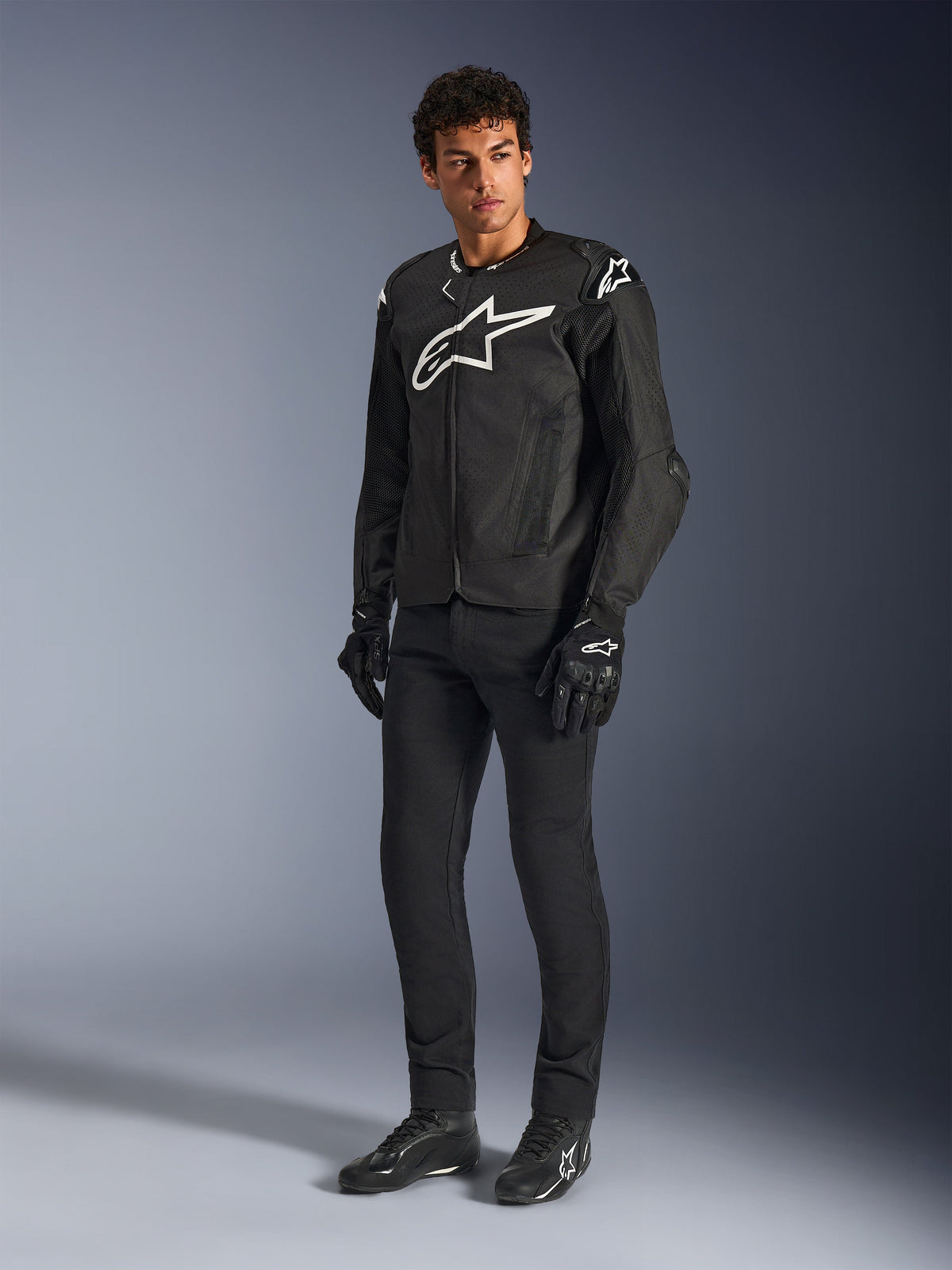 Alpinestars T-GP Air Jacke, schwarze Textil-Motorradjacke, getragen von einem Model mit passender schwarzer Hose und Handschuhen, markantes weißes Alpinestars-Logo auf Brust und Schultern, belüftete Mesh-Paneele, sportliche, vom Rennsport abgeleitete Passform