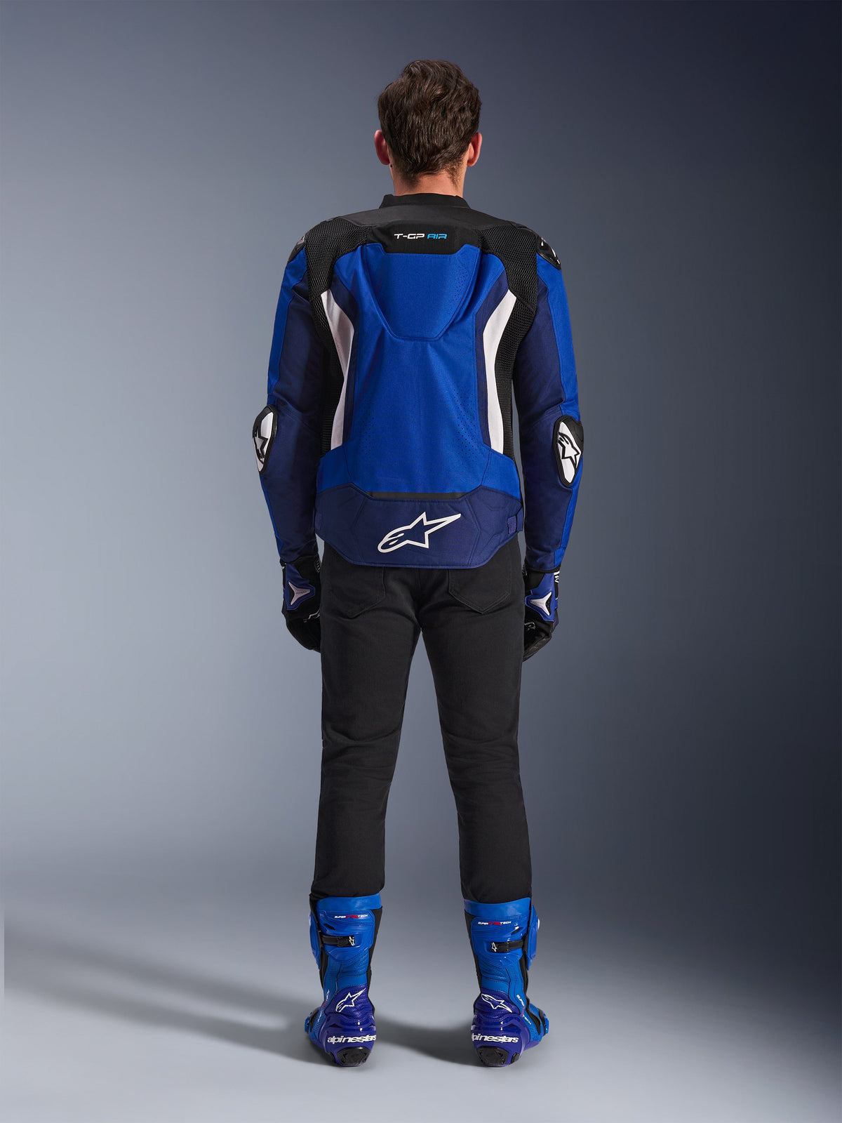 Alpinestars T-GP Air Jacke, Hellblau Navy Weiß, Rückansicht mit aerodynamischem Höcker und Mesh-Belüftungseinsätzen, weißes Alpinestars-Logo am unteren Rücken, kombiniert mit schwarzer Hose und blauen Rennstiefeln.