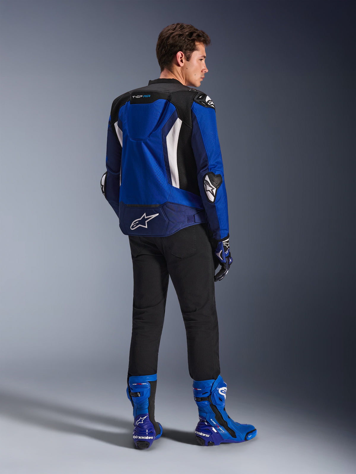 Alpinestars T-GP Air Jacke, Sport-Textiljacke, Hellblau, Marineblau und Weiß, Rückansicht an einem Model in schwarzer Hose und passenden blau-weißen Stiefeln, mit weißem Alpinestars-Logo am unteren Rücken, Mesh-Belüftungseinsätzen und einem aerodynamischen Höcker