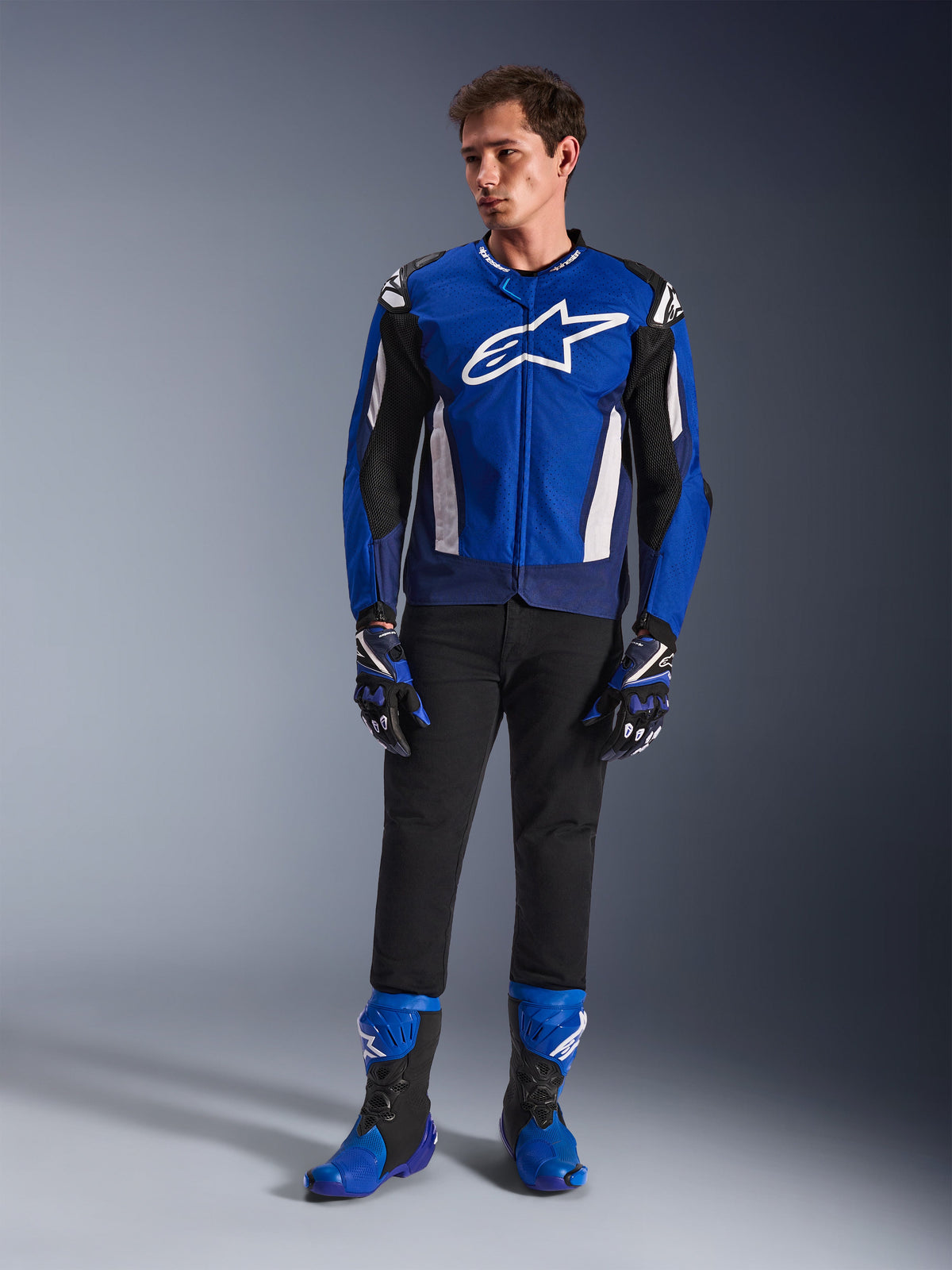 Männliches Model in der Alpinestars T-GP Air Jacke in Bright Blue Navy White, kombiniert mit einer schwarzen Hose, passenden blau-dunkelblauen Rennhandschuhen und blau-schwarzen Motorradstiefeln, Studioaufnahme eines kompletten Sport-Fahroutfits.
