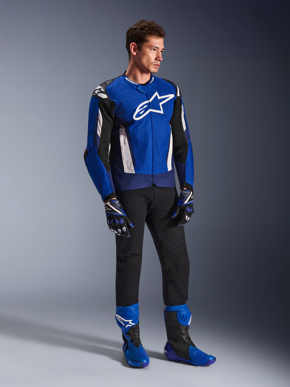 Alpinestars T-GP Air Jacke, sportliche Textil-Motorradjacke, Bright Blue Navy und Weiß, getragen mit passenden blau-schwarzen Handschuhen, einer schwarzen Hose und blauen Rennstiefeln, mit weißem Logo auf der Brust und technischen Mesh-Einsätzen, Ganzkörper-Studioaufnahme
