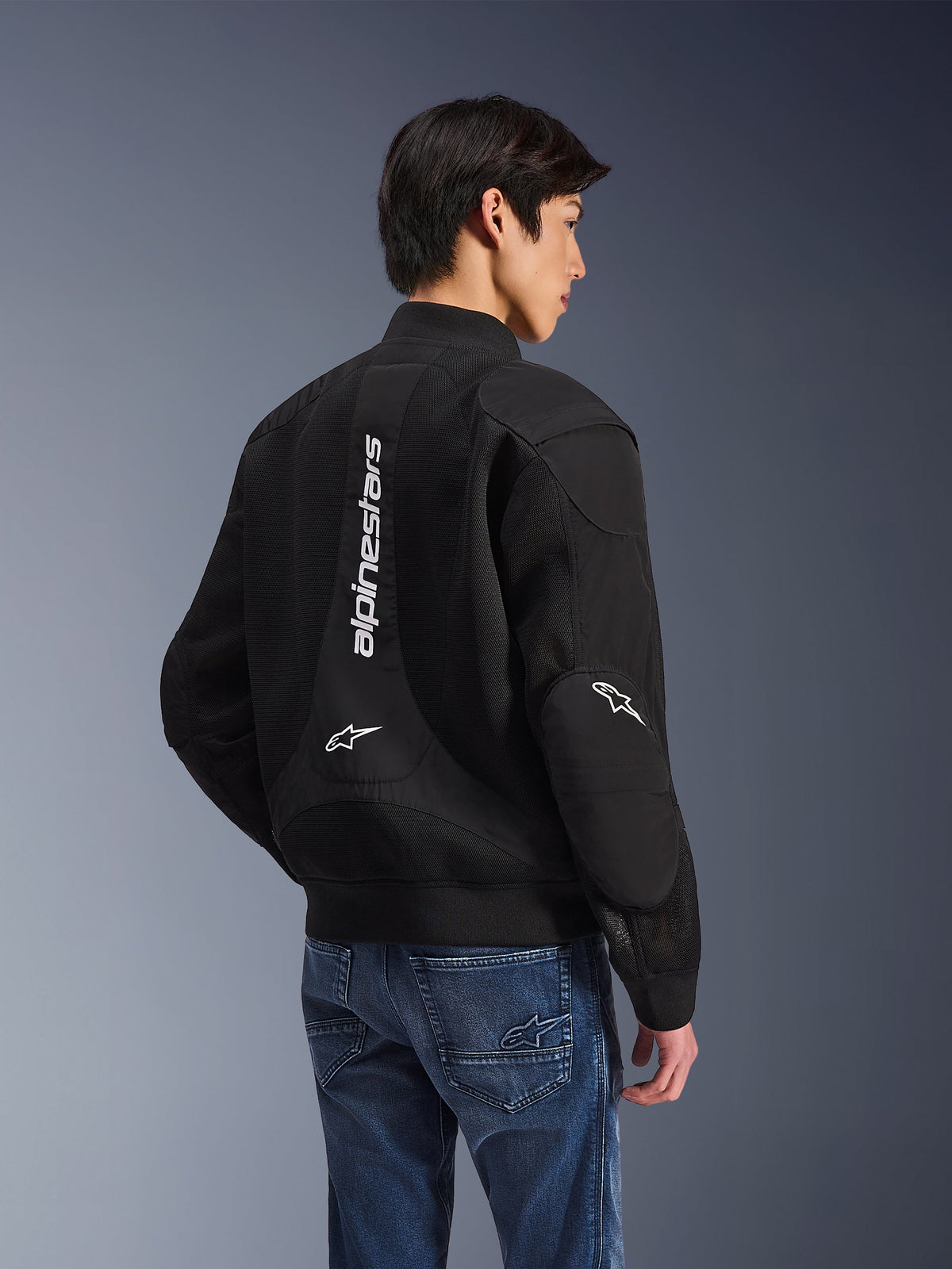 Alpinestars Flight Air Jacke, schwarze Motorradjacke, Rückansicht mit großem vertikalem weißem Logo und Mesh-Belüftungseinsätzen, Bomber-Stil mit geripptem Saum, getragen mit blauen Jeans, urbane zeitgenössische Motorradbekleidung