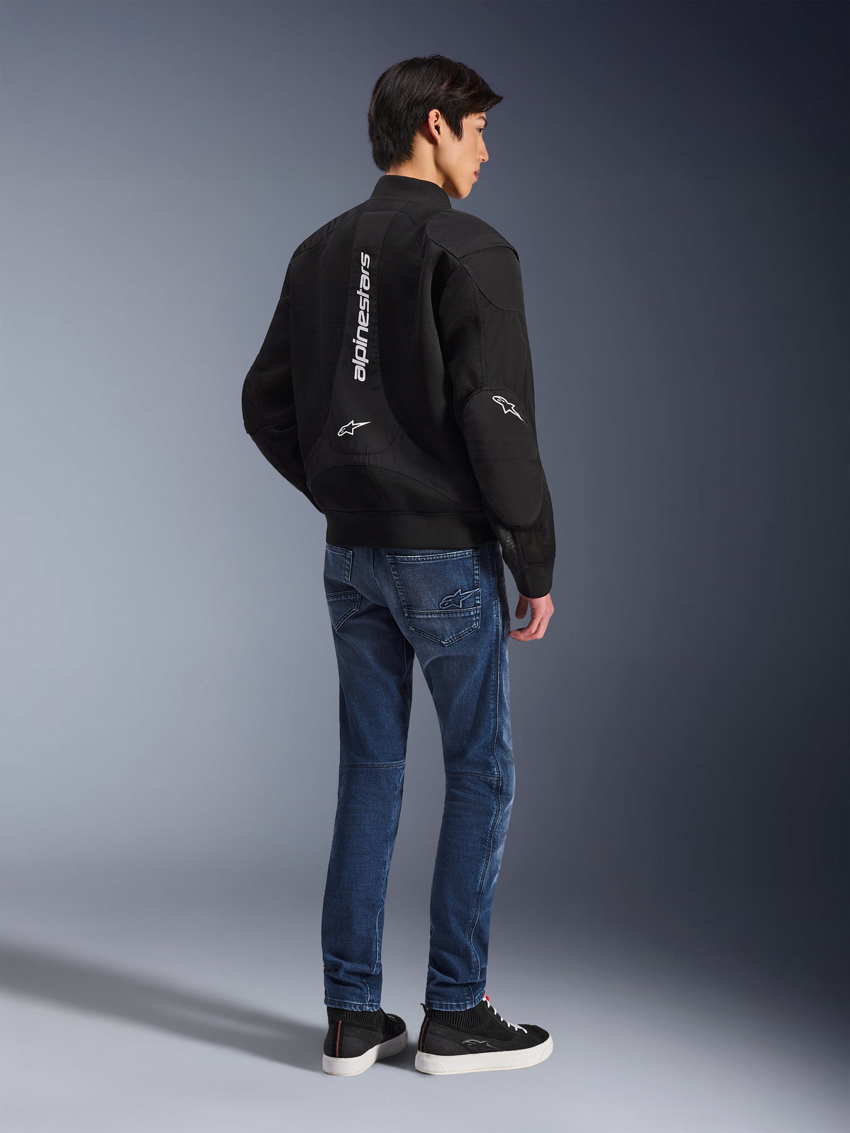 Model in der Alpinestars Flight Air Jacke in Schwarz, Rückansicht, kombiniert mit blauen Motorradjeans und schwarzen Sneakern, mit vertikalem weißem Alpinestars-Schriftzug und 3D-Mesh-Belüftungseinsätzen.