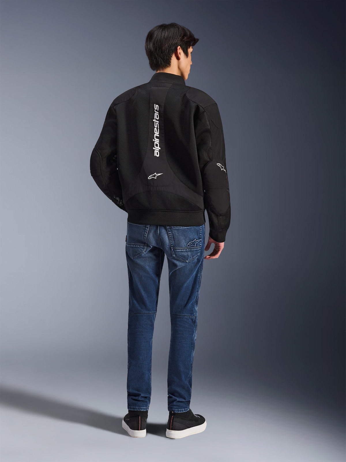 Model trägt die Alpinestars Flight Air Jacke von hinten, schwarze Motorradjacke im Bomber-Stil mit 3D-Mesh-Paneelen und vertikalem weißem Logo, kombiniert mit blauen Denim-Fahrjeans und schwarzen Sneakern, moderne urbane Motorradausrüstung