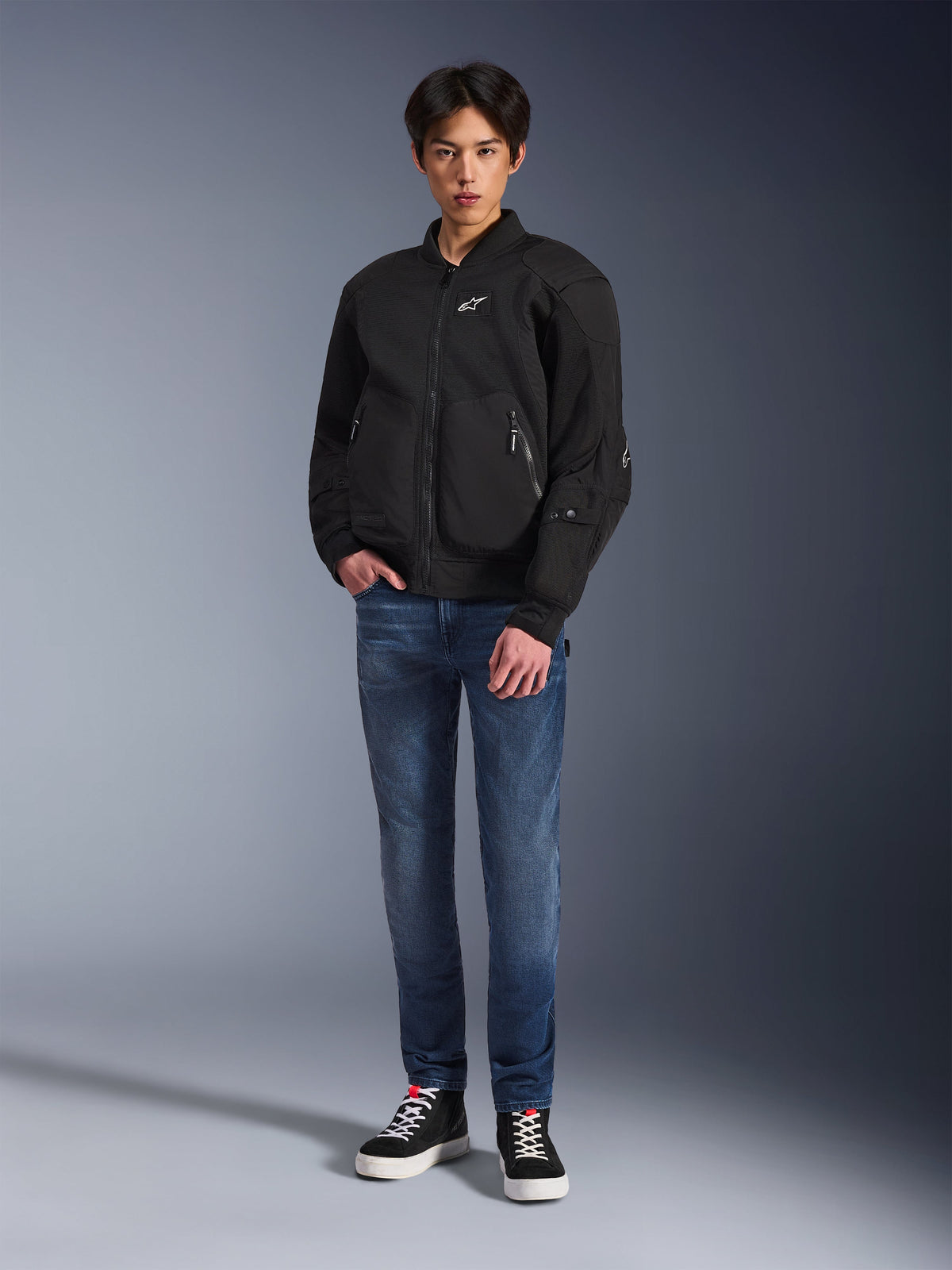 Model in der Alpinestars Flight Air Jacke in Schwarz, eine urbane Motorradjacke im Bomber-Stil mit 3D-Mesh-Einsätzen und Frontreißverschluss, kombiniert mit blauen Jeans und schwarzen Sneakern, moderne Streetwear-inspirierte Motorradbekleidung.
