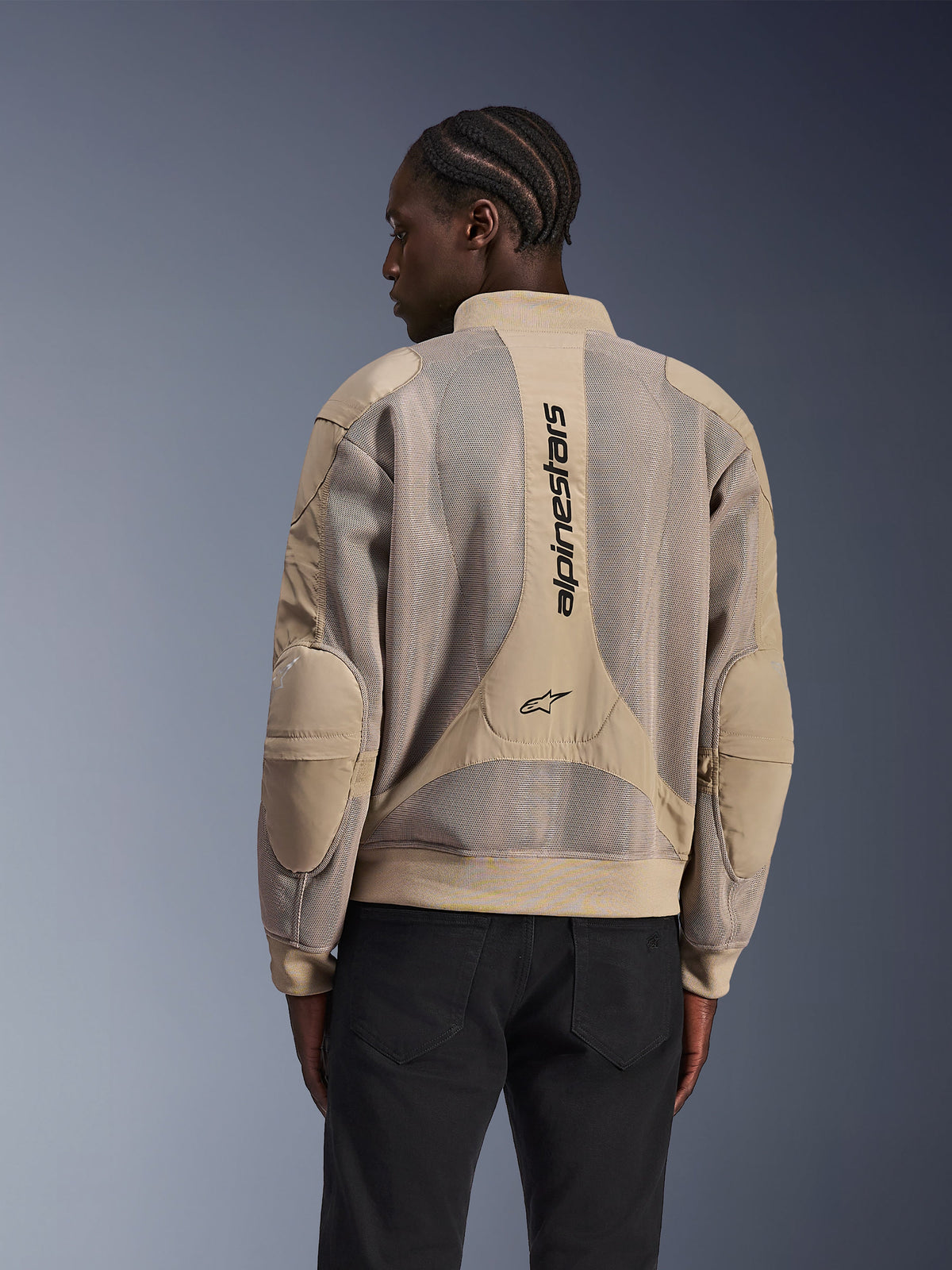 Alpinestars Flight Air Jacke, urbane Motorradjacke, Tan, Rückansicht mit erweiterten 3D-Mesh-Paneelen und vertikalem Alpinestars-Branding auf dem Rücken, Bomberjacken-inspiriertes Design mit geripptem Saum, getragen von einem Model in schwarzer Hose