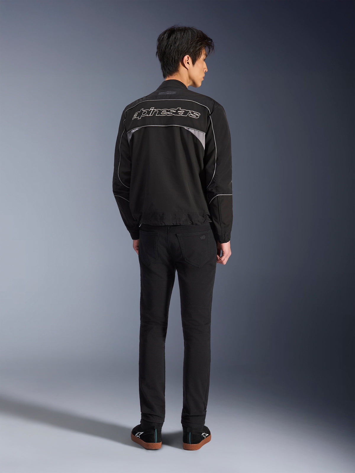 Alpinestars Unite Jacke, Motorradjacke in Schwarz und Dunkelgrau, Rückansicht mit Alpinestars-Umrisslogo auf dem Rücken, graue reflektierende Einsätze, minimalistisches urbanes Design, getragen mit schwarzer Hose und schwarzen Schuhen.