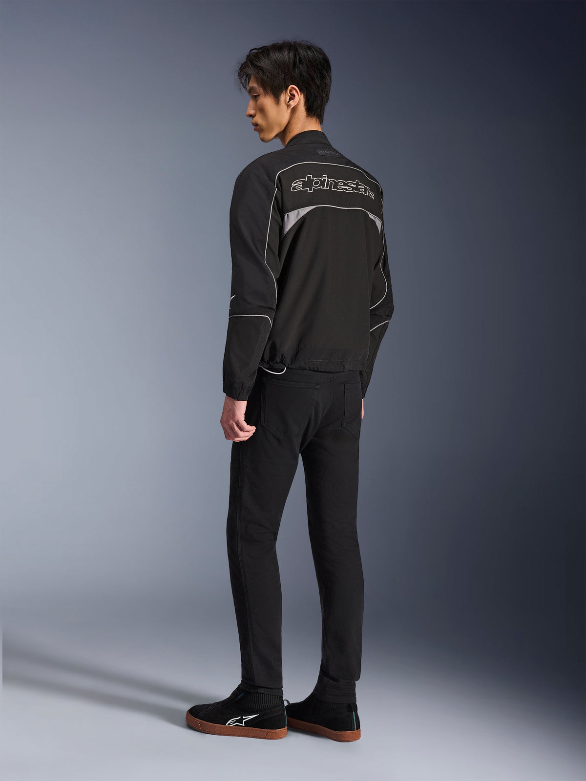 Alpinestars Unite Jacke, Urban-Motorradjacke, Schwarz und Dunkelgrau, Rückansicht mit großem umrandetem Alpinestars-Logo, reflektierende Paspeln und Einsätze, minimalistisches Softshell-Design, getragen mit schwarzer Hose und Freizeitschuhen