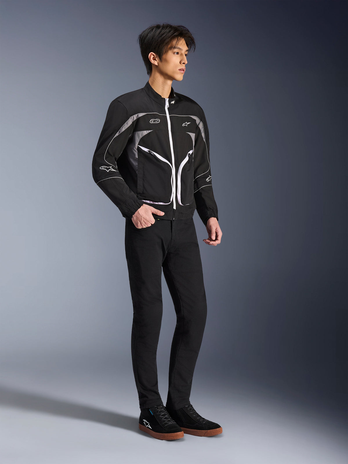 Model in der Alpinestars Unite Jacke in Schwarz und Dunkelgrau, urbane Motorradjacke mit weißen Reißverschlüssen und reflektierenden Details, kombiniert mit einer schwarzen Slim-Fit-Hose und Sneakern, minimalistische Streetwear-inspirierte Fahrausrüstung.