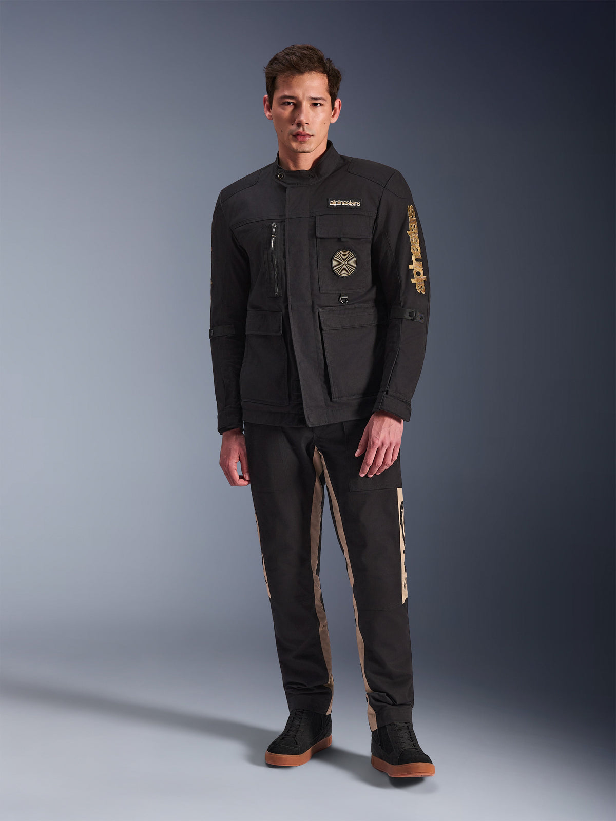 Male model wearing the Alpinestars Dice Jacket, Black urban motorcycle jacket with vintage field jacket styling, four functional front pockets, Alpinestars branding on the sleeve and chest, paired with black and tan pants, modern urban riding gear