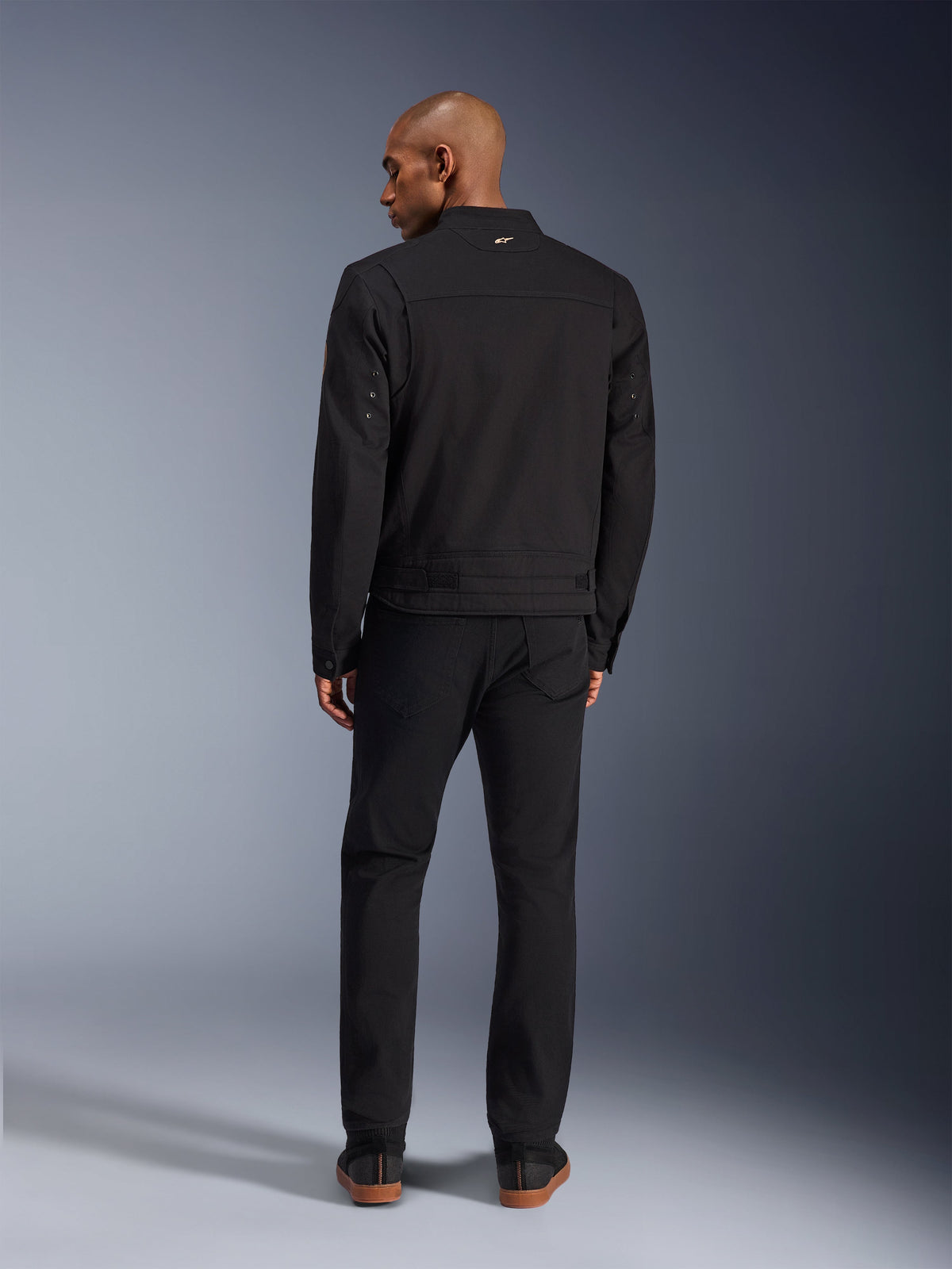 Alpinestars Bruiser Jacke in Schwarz, Rückansicht eines männlichen Models in der urbanen Motorradjacke mit passender schwarzer Hose, mit kleinem goldenem Logo am Kragen und verstellbaren Taillenriemen, von Workwear inspirierte Ästhetik