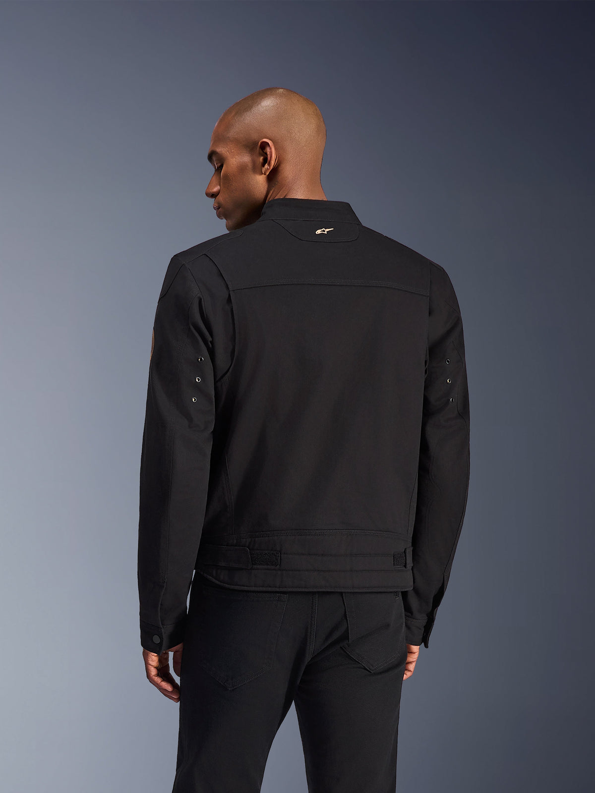 Alpinestars Bruiser Jacke, schwarze Urban-Motorradjacke, Rückansicht, mit einem von Workwear inspirierten Design, einem kleinen goldenen Logo am Kragen und verstellbaren Taillenriegeln, strapazierfähige Textilkonstruktion für urbane Fahrten