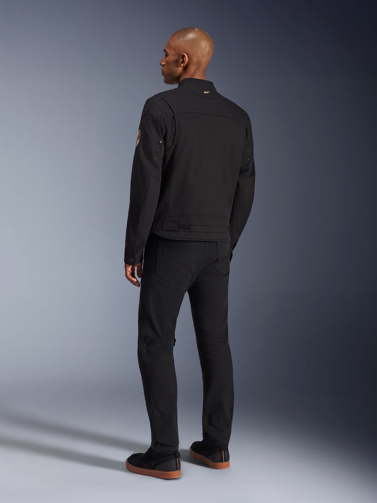 Alpinestars Bruiser Jacke, urbane schwarze Motorradjacke, Rückansicht am Modell, klassisches Workwear-Styling, mit plissierten Schulterpaneelen und Oscar-Spiral-Aufnäher am Ärmel, kombiniert mit schwarzer Hose und lässigen schwarzen Schuhen mit Gummisohlen.