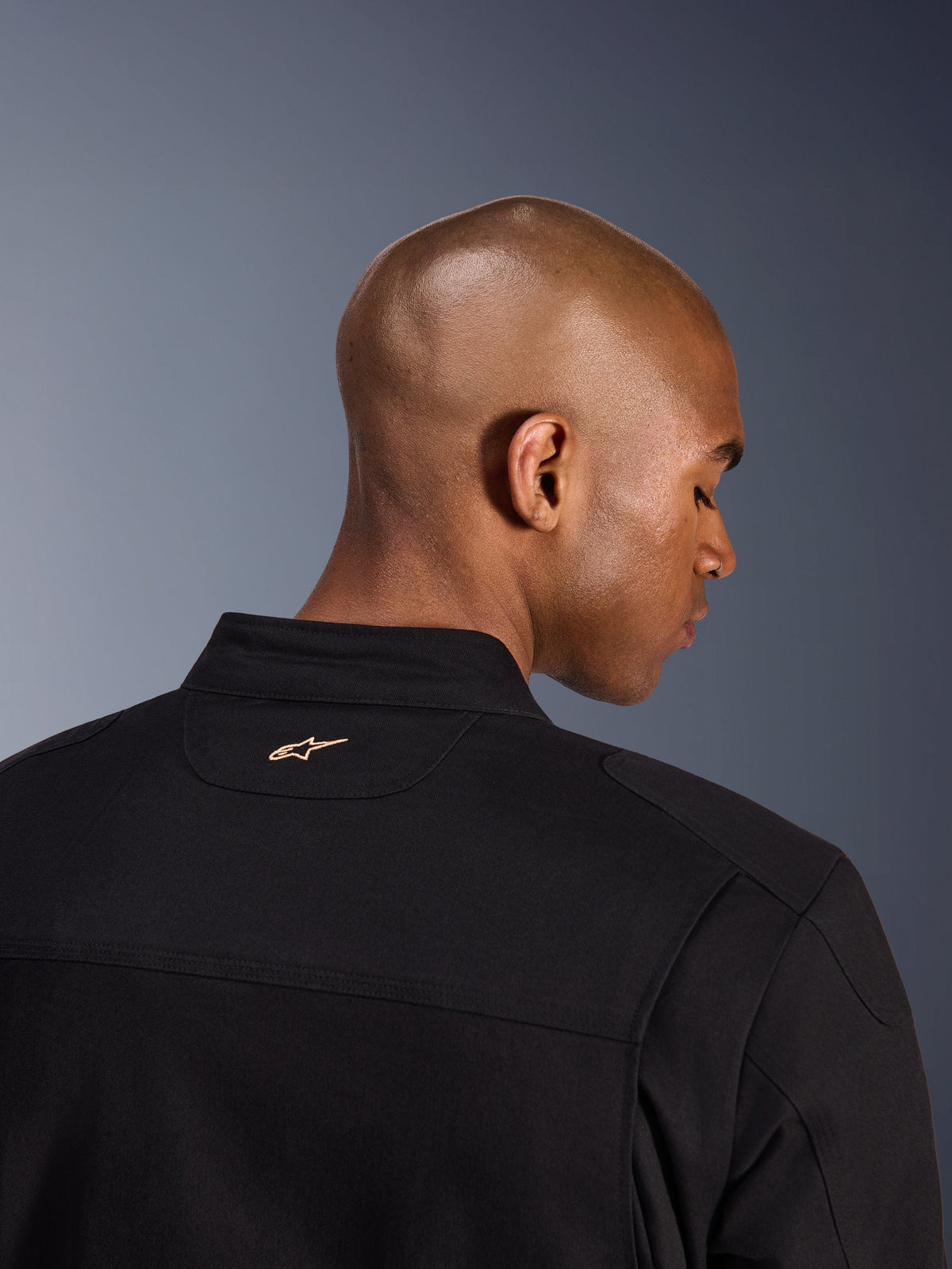 Alpinestars Bruiser Jacke, Schwarz, Detailaufnahme der Rückseite von Kragen und Schultern, kleines hellbraun besticktes Alpinestars-Logo im Nacken, urbane Motorradjacke mit Workwear-Ästhetik