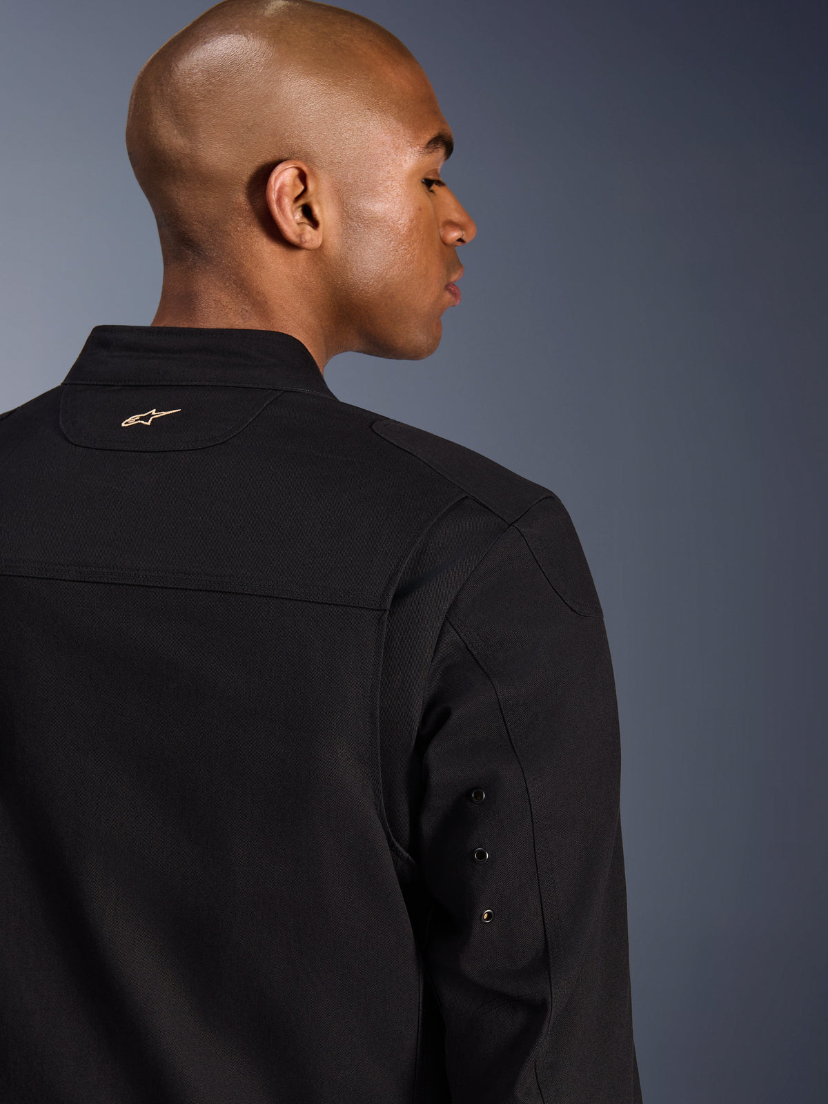 Alpinestars Bruiser Jacke, textile urbane Motorradjacke, Schwarz, Nahaufnahme der Rückansicht mit besticktem goldenem Logo am oberen Rücken, Metallösen-Details am Ärmel und plissierten Schulterbahnen für verbesserte Bewegungsfreiheit.