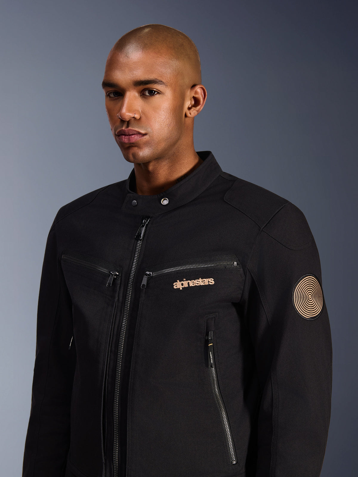 Männliches Model in Alpinestars Bruiser Jacke, Schwarz, urbane Motorradjacke aus Polycotton-Twill, mit Reißverschlusstaschen, goldfarben besticktem Logo auf der Brust und Oscar-Spiral-Aufnäher am Ärmel, moderner Pendler-Stil