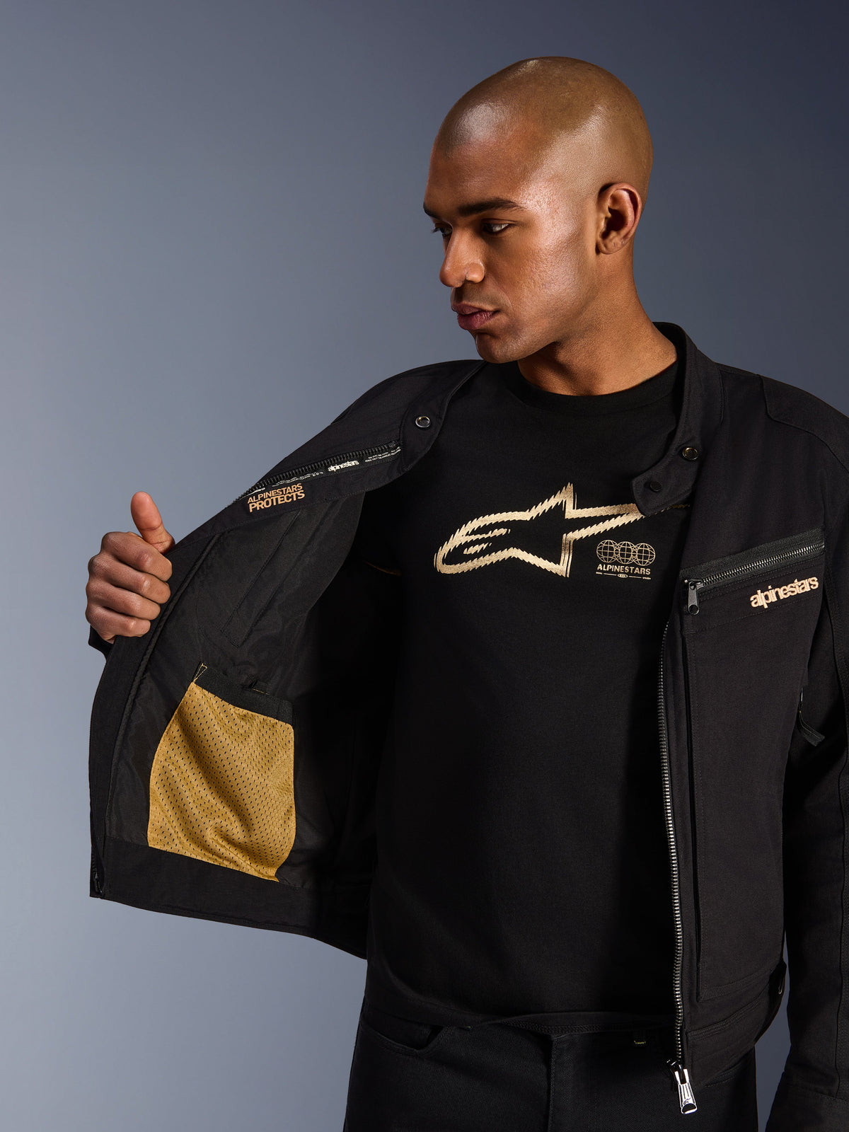 Alpinestars Bruiser Jacke, schwarze urbane Motorradjacke, Nahaufnahme der geöffneten Innenseite mit goldener Mesh-Tasche und „Alpinestars Protects“-Branding, getragen über einem schwarzen Logo-T-Shirt, textile Fahrerausrüstung mit Workwear-Ästhetik
