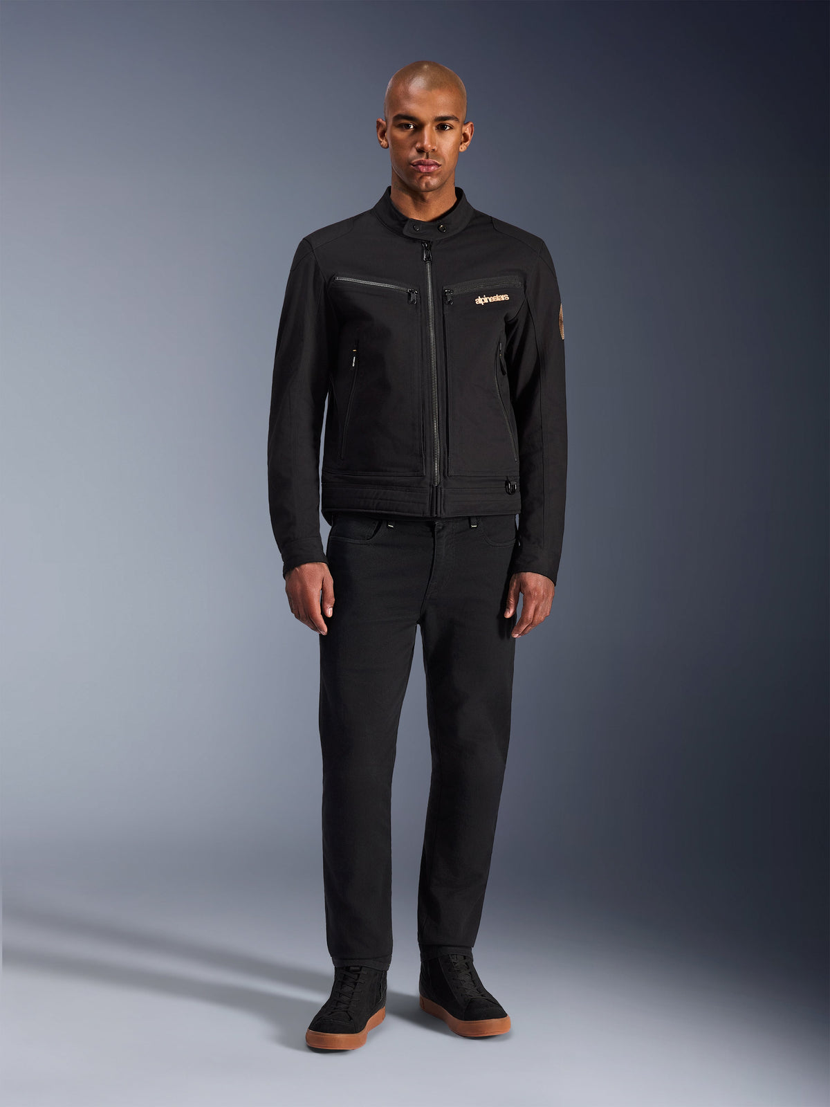 Alpinestars Bruiser Jacke, schwarze urbane Motorradjacke, getragen von einem Model mit schwarzer Hose und High-Top-Sneakern, Vorderansicht mit Brusttaschen mit Reißverschluss und besticktem Logo, klassisches, von Workwear inspiriertes Design für den täglichen Weg zur Arbeit