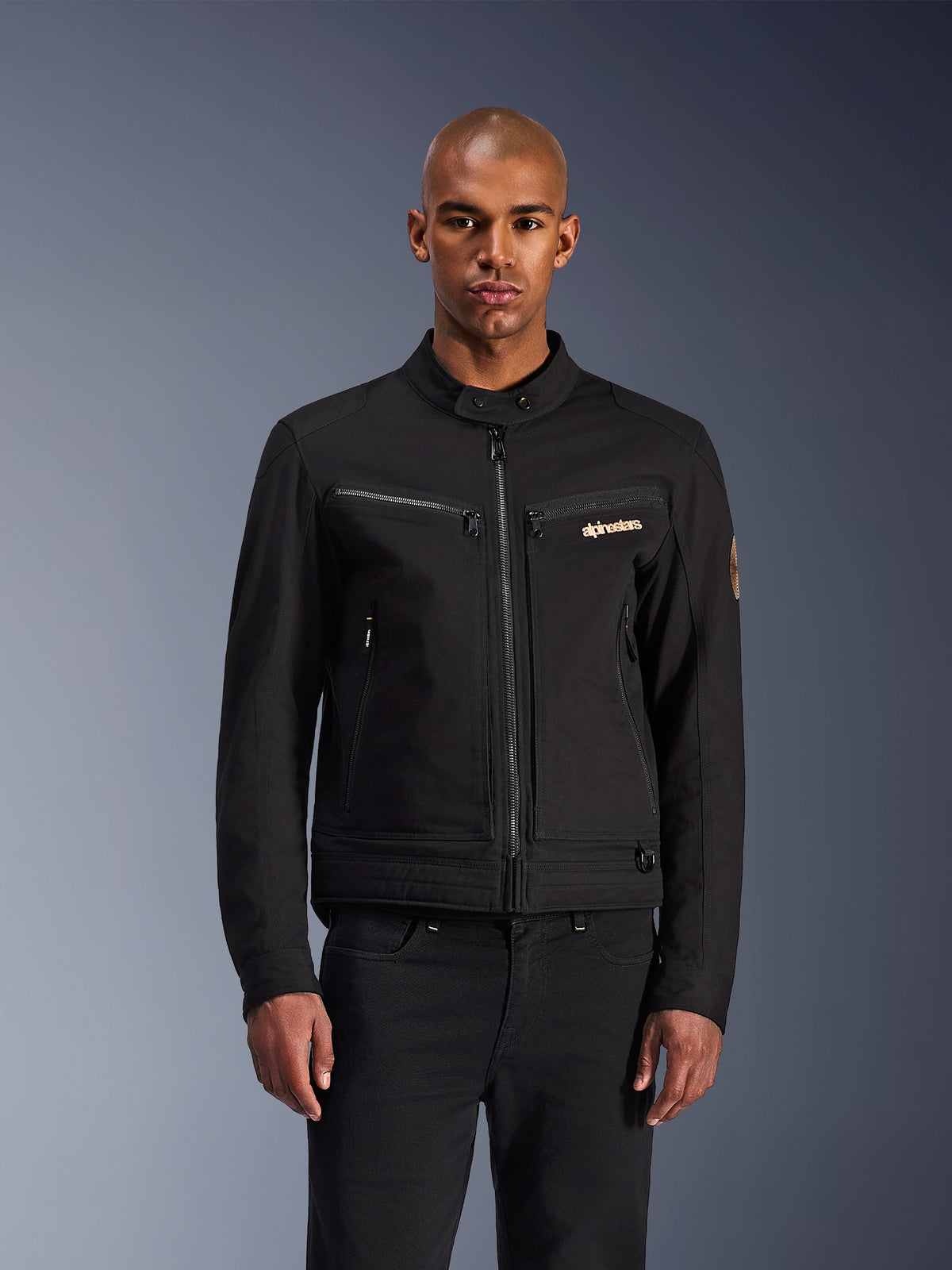 Alpinestars Bruiser Jacke, Schwarze urbane Motorradjacke, Frontansicht an einem Model, klassische Workwear-Ästhetik, Brusttaschen mit Reißverschluss, Stehkragen mit Druckknopf und Alpinestars-Logo, ideal für urbane Fahrer