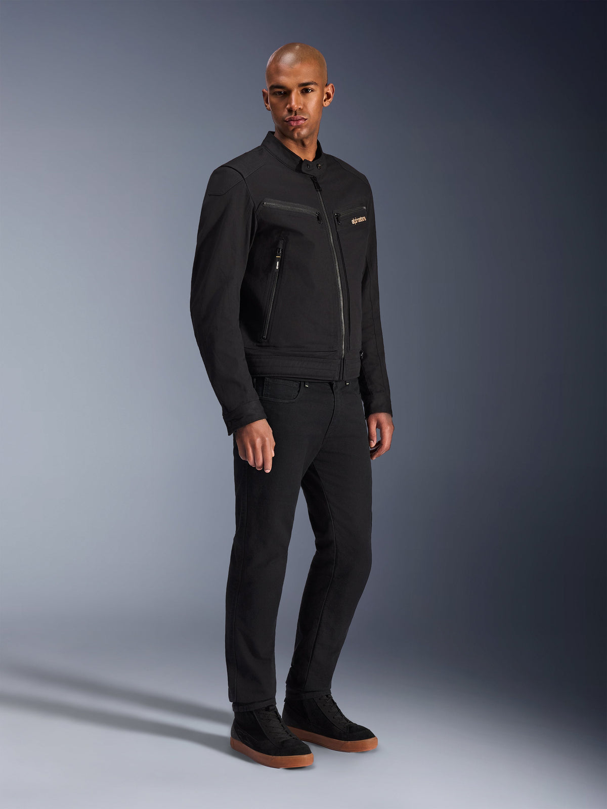 Alpinestars Bruiser Jacke, urbane Motorradjacke, Schwarz, getragen von einem Modell mit schwarzer Hose und Sneakern, mit Brusttaschen mit Reißverschluss, Polycotton-Twill-Gewebe und einem eleganten, von Workwear inspirierten Design für urbanes Fahren