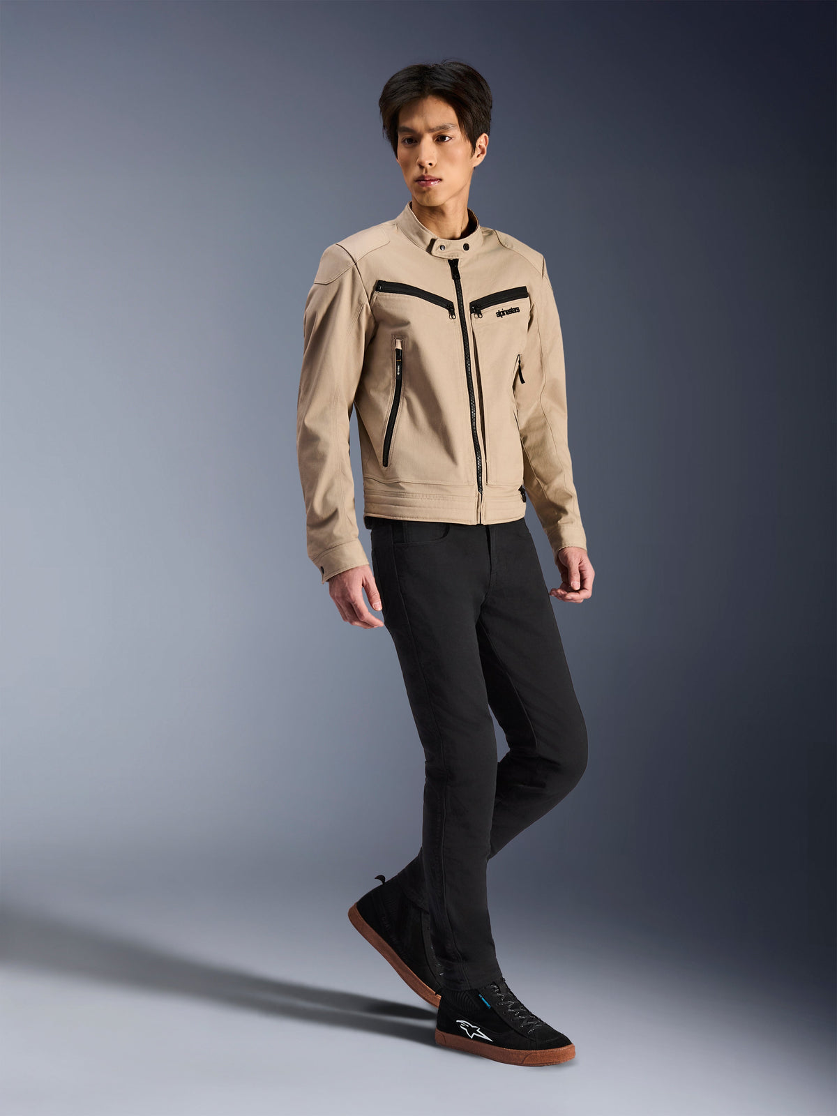 Alpinestars Bruiser Jacket, Urban Motorcycle Jacket, Tan, worn by a male model with black slim-fit pants and black sneakers, workwear-inspired textile construction with black zipper accents, contemporary urban street style.