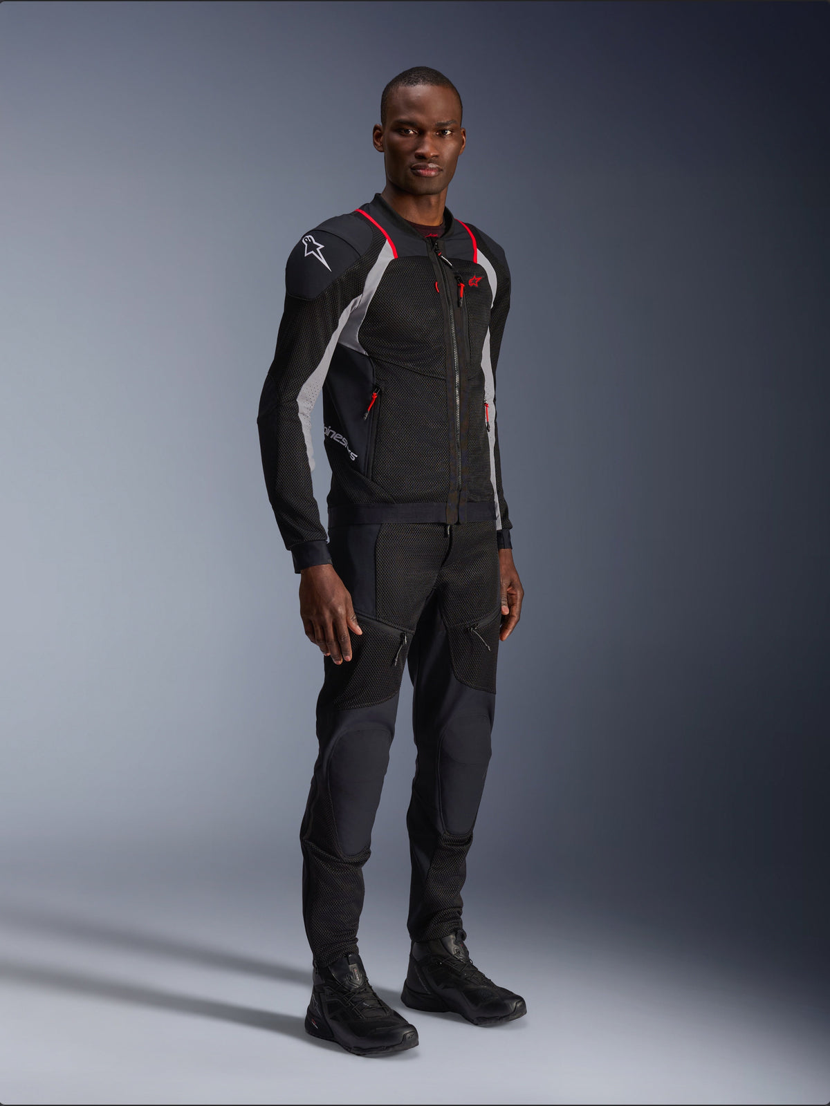 Alpinestars Troop-Air Pro Jacke, Schwarz, Ash Gray und Bright Red, technische Mesh-Protektorenjacke, mit roten Akzenten und weißem Logo auf der Schulter, getragen mit passender schwarzer Hose und Stiefeln, moderne urbane Motorradbekleidung