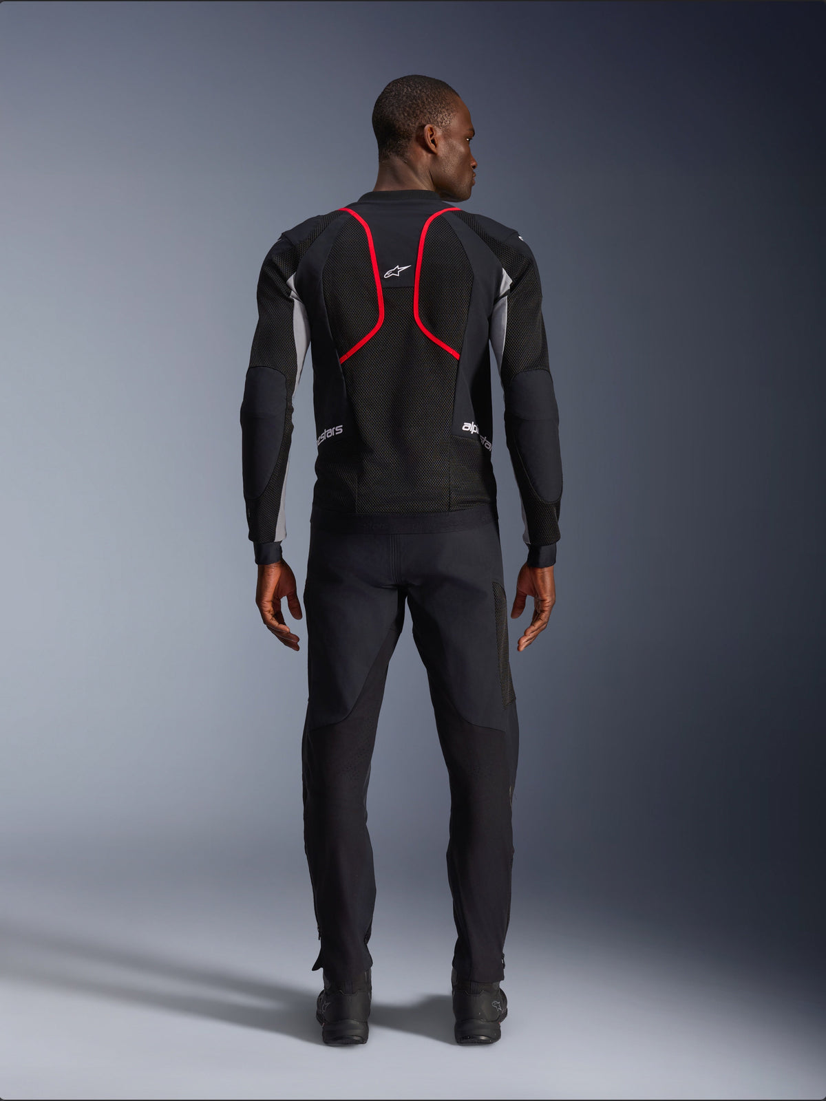 Modell in der Rückansicht in der Alpinestars Troop-Air Pro Jacke, Schwarz mit aschgrauen und leuchtend roten Akzenten, technische Mesh-Konstruktion mit markanten roten Paspeln am Rücken, Alpinestars-Logo im oberen Rückenbereich, kombiniert mit einer schwarzen Motorradhose, hochwertige gepanzerte Air-Jacke für den Straßeneinsatz