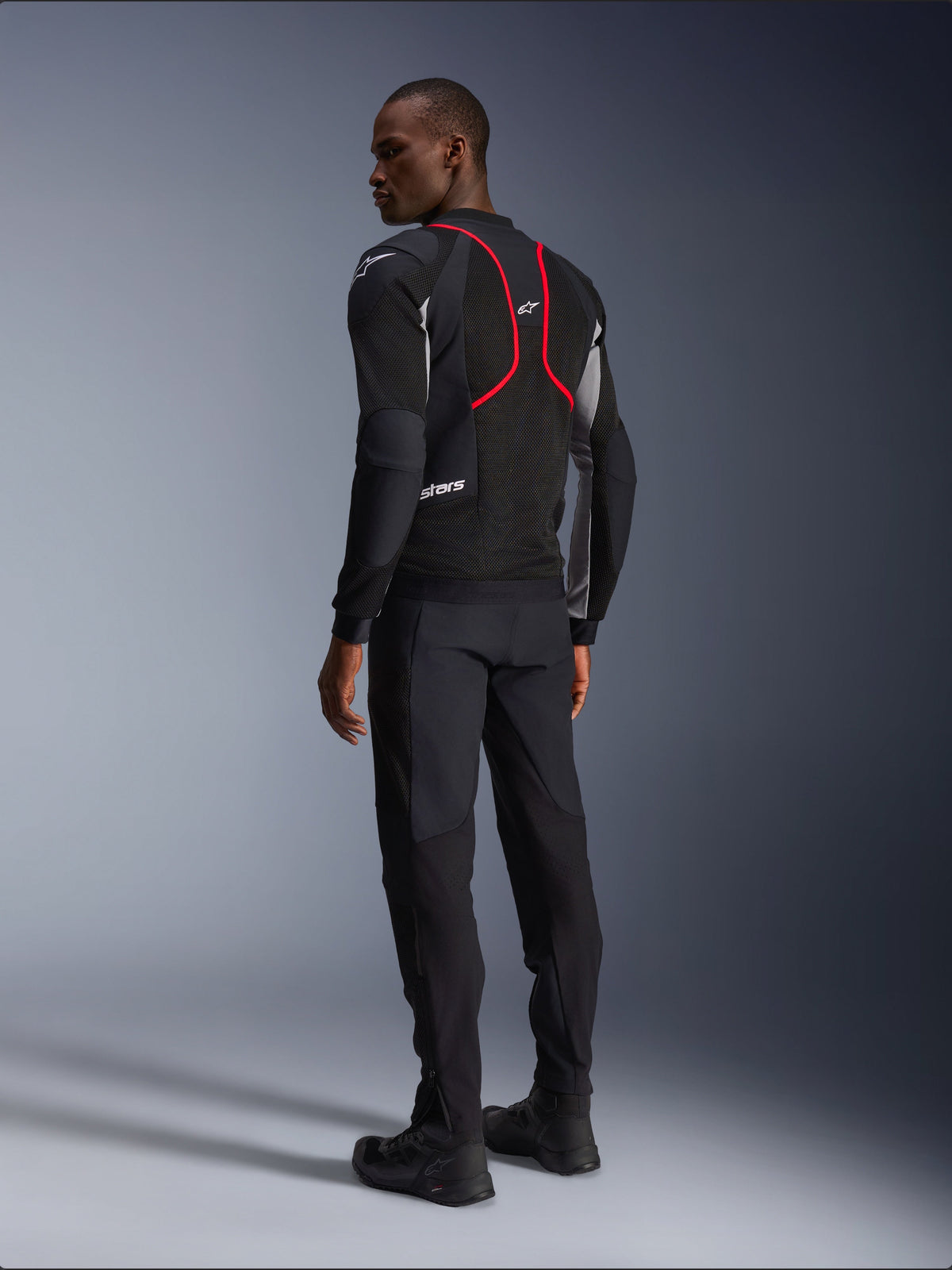 Alpinestars Troop-Air Pro Jacke, Motorradjacke, Schwarz, Eschegrau und Hellrot, Rückansicht am Modell, mit roten Akzentlinien und Mesh-Belüftungseinsätzen, getragen mit schwarzer Hose und Stiefeln, urbaner Motorradstil