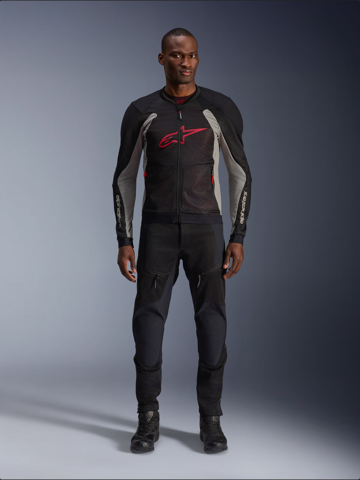 Male model wearing the Alpinestars Troop-Air Jacket in Black Ash Gray and Bright Red, technical mesh motorcycle jacket featuring a prominent red chest logo, styled with black motorcycle pants and boots, urban road riding apparel