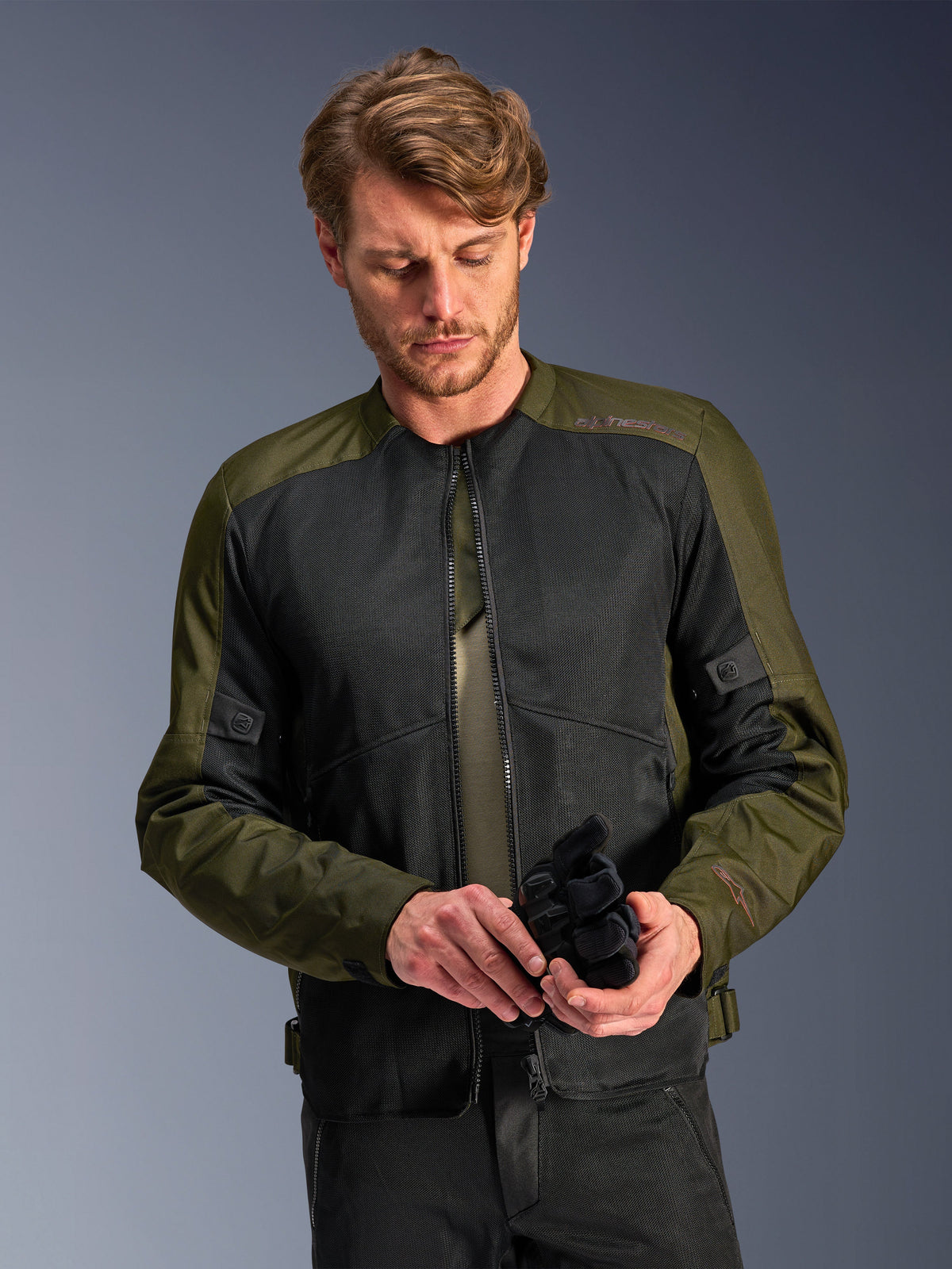 Mann in der Alpinestars C-1 Air Jacke in Khaki, urbane Motorradjacke mit olivgrünem Textil und schwarzen Mesh-Einsätzen, blickt nach unten und hält schwarze Motorradhandschuhe, cleaner und zeitgemäßer Stil für Straßenfahrten.