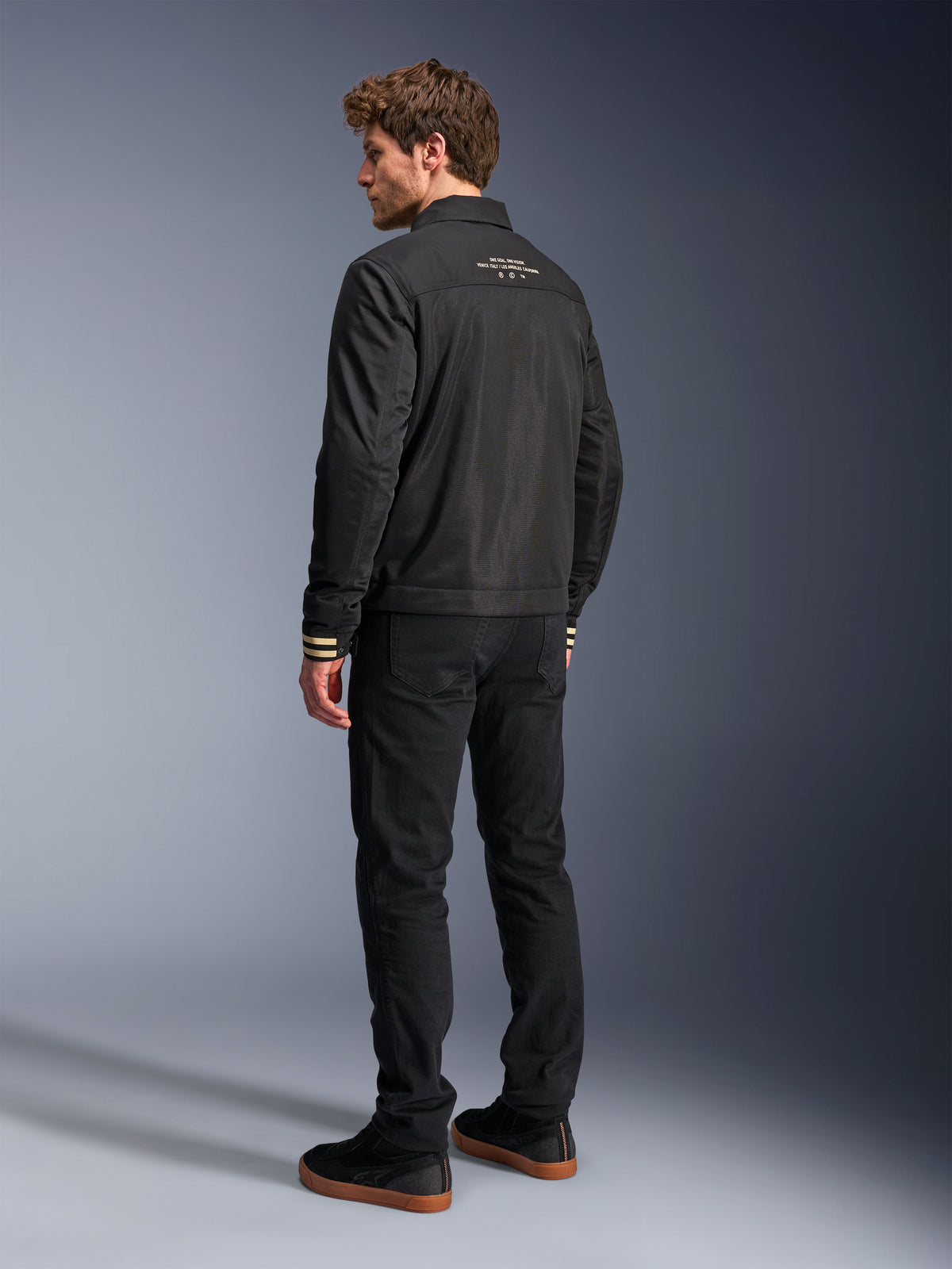 Alpinestars Ignite Air Jacket, Black motorcycle jacket, rear view featuring white graphic text on the upper back and tan striped cuffs, Harrington-style silhouette with mesh body, paired with black pants and black sneakers