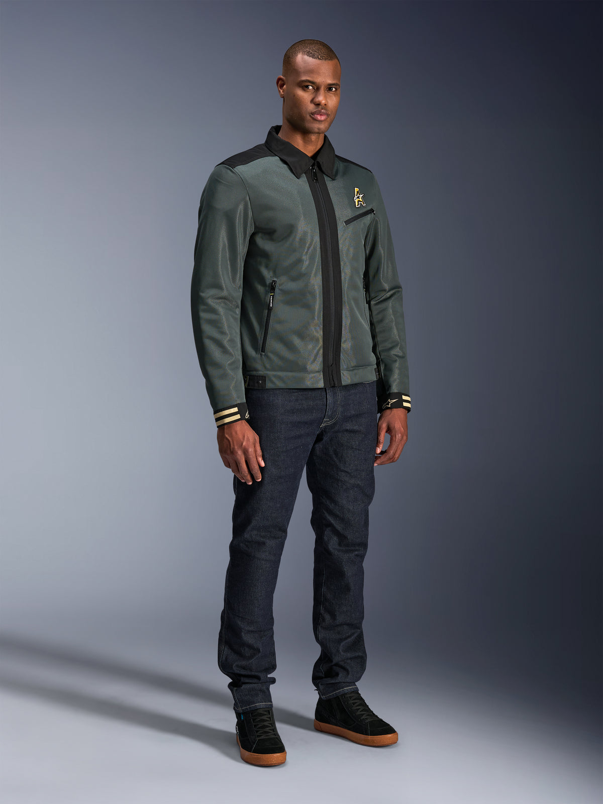 Alpinestars Ignite Air Jacke, Urban Gray Black Blue, urbane Motorradjacke, getragen von einem Model mit dunkler Denim-Jeans und schwarzen Sneakern, mit Mesh-Hauptteil, traditionellem Hemdkragen, Harrington-inspirierter Silhouette und goldenem Alpinestars-Branding.