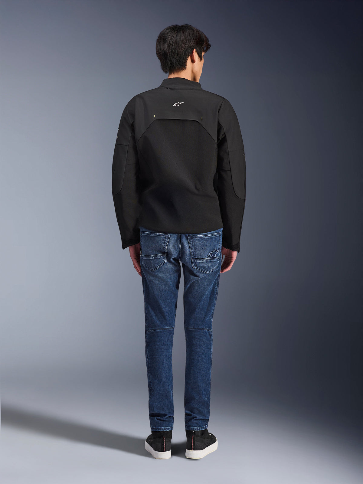 Rückansicht eines Modells in der Alpinestars Provoke Jacke, schwarze technische Textil-Motorradjacke, kombiniert mit blauen Denim-Motorradjeans und schwarzen Sneakern, zeigt die Rückenkonstruktion und das kleine weiße Alpinestars-Logo, urbanes zeitgemäßes Design