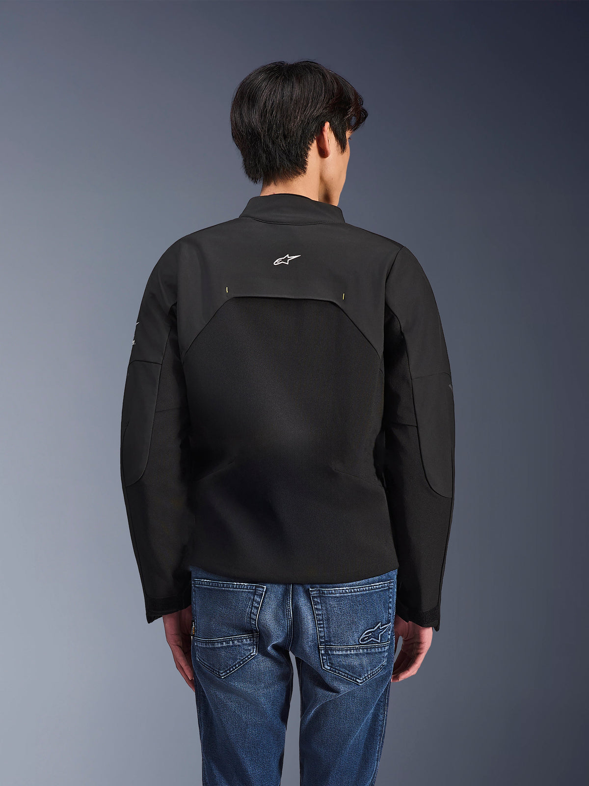 Alpinestars Provoke Jacke, Urbane Motorradjacke, Schwarz, Rückansicht mit weißem Astars-Logo und technischen Ripstop-Einsätzen, kombiniert mit blauen Denim-Jeans, modernes Design für den Stadtverkehr