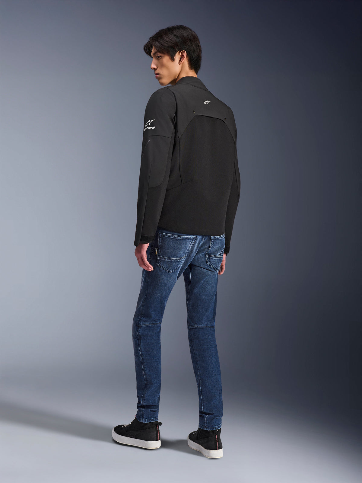 Alpinestars Provoke Jacke, schwarze urbane Motorradjacke, getragen von einem Model in einer Dreiviertel-Rückansicht, kombiniert mit blauen Denim-Jeans und schwarzen Sneakern, mit Ripstop-Softshell-Einsätzen und weißem Alpinestars-Branding auf dem Ärmel und dem Rücken.