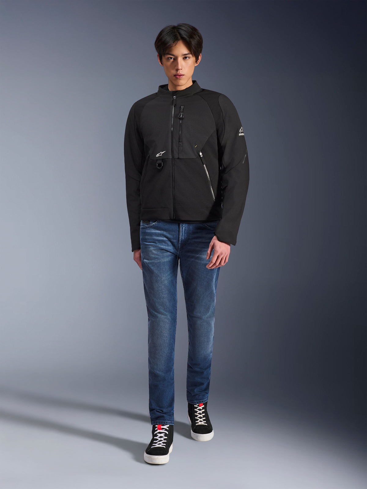 Männliches Model in der Alpinestars Provoke Jacke in Schwarz, urbane Textil-Motorradjacke mit technischen Reißverschlüssen und Ripstop-Softshell-Einsätzen, kombiniert mit blauen Jeans und schwarzen Sneakern, moderner urbaner Rider-Stil.