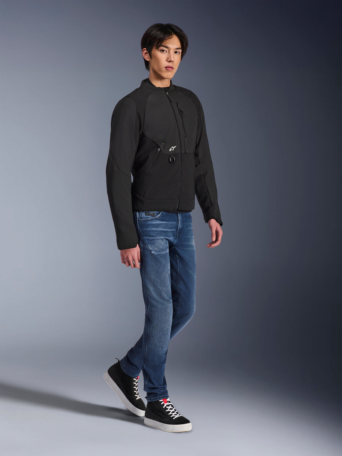 Alpinestars Provoke Jacke, Urbane Motorradjacke, Schwarz, getragen von einem männlichen Model mit blauen Jeans und schwarzen Sneakern, technisches Textildesign mit kleinem Alpinestars-Logo, Ganzkörper-Lifestyle-Aufnahme für zeitgenössisches Fahren in der Stadt