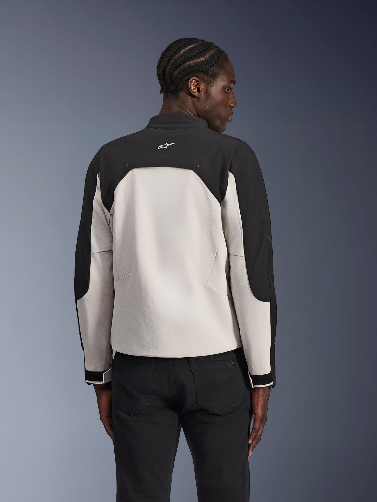 Alpinestars Provoke Jacke, Urbane Motorradjacke, Black Metal Gray, Rückansicht an einem Model, zweifarbiges Textildesign mit schwarzen Schultern und hellgrauem Rückenpanel, Alpinestars Stern-Logo am hinteren Kragen, kombiniert mit schwarzer Hose vor einem blauen Hintergrund mit Farbverlauf