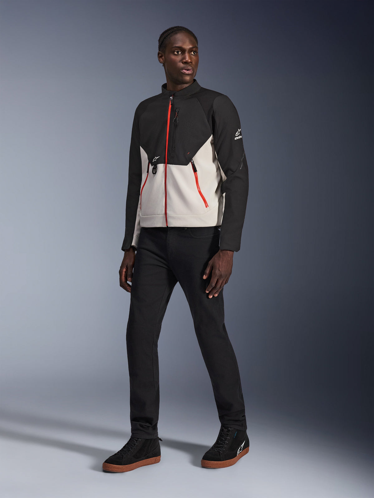 Modell trägt die Alpinestars Provoke Jacke in Black Metal Gray, mit markanten roten Front- und Taschenreißverschlüssen, kombiniert mit einer schwarzen Hose und schwarzen High-Top-Sneakern mit Gummisohlen, urbane zeitgenössische Motorradausrüstung für einen straßentauglichen Look.