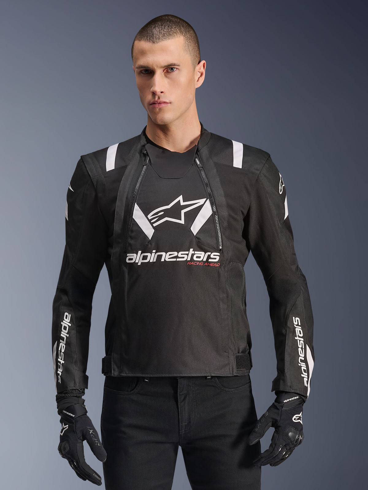 Alpinestars T-Stunt Air Jacke, schwarz-weiße Motorradjacke, getragen von einem Mann mit schwarzen Handschuhen und Hosen, mit markantem weißem Branding auf der Brust und den Ärmeln sowie roten „Racing Ahead“-Akzenten, sportliches Design mit weißen Schulterstreifen und atmungsaktiven Mesh-Einsätzen.