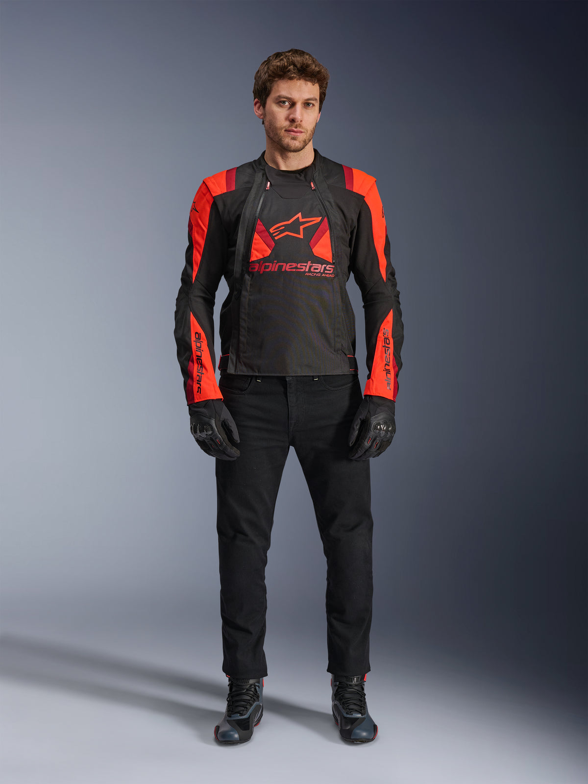 Alpinestars T-Stunt Air Jacke, Schwarz, Leuchtrot und Dunkelrot, getragen von einem Model mit schwarzen Motorradjeans und Handschuhen, mit modularem 2-in-1-Design und markanten Mesh-Einsätzen für Luftzirkulation, sportlich geschnittene Motorradausrüstung für die Straße