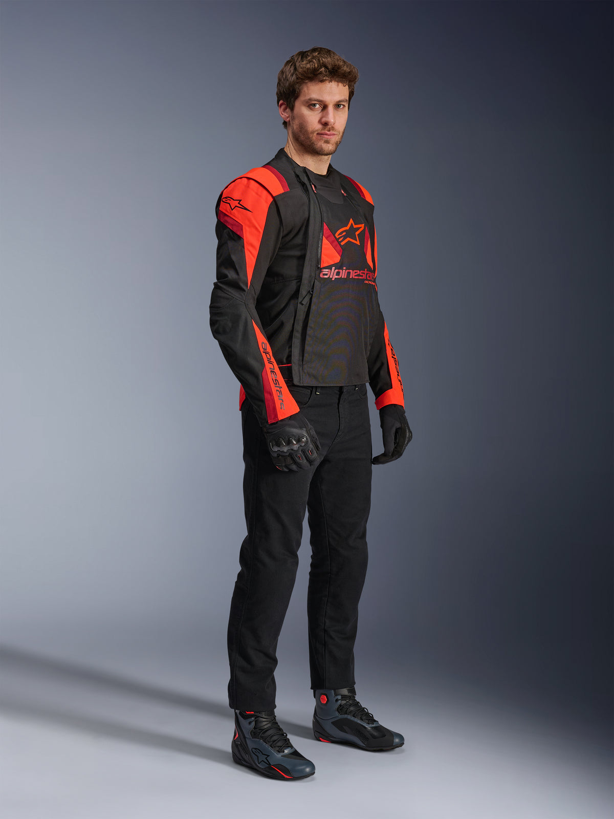 Alpinestars T-Stunt Air Jacke, Schwarz, Red Fluo und Dunkelrot, modulare Motorradjacke, von einem Model getragen mit geöffneten Frontpanels zur Freigabe des Mesh-Innenfutters, kombiniert mit schwarzer Fahrhose, Handschuhen und Schuhen.