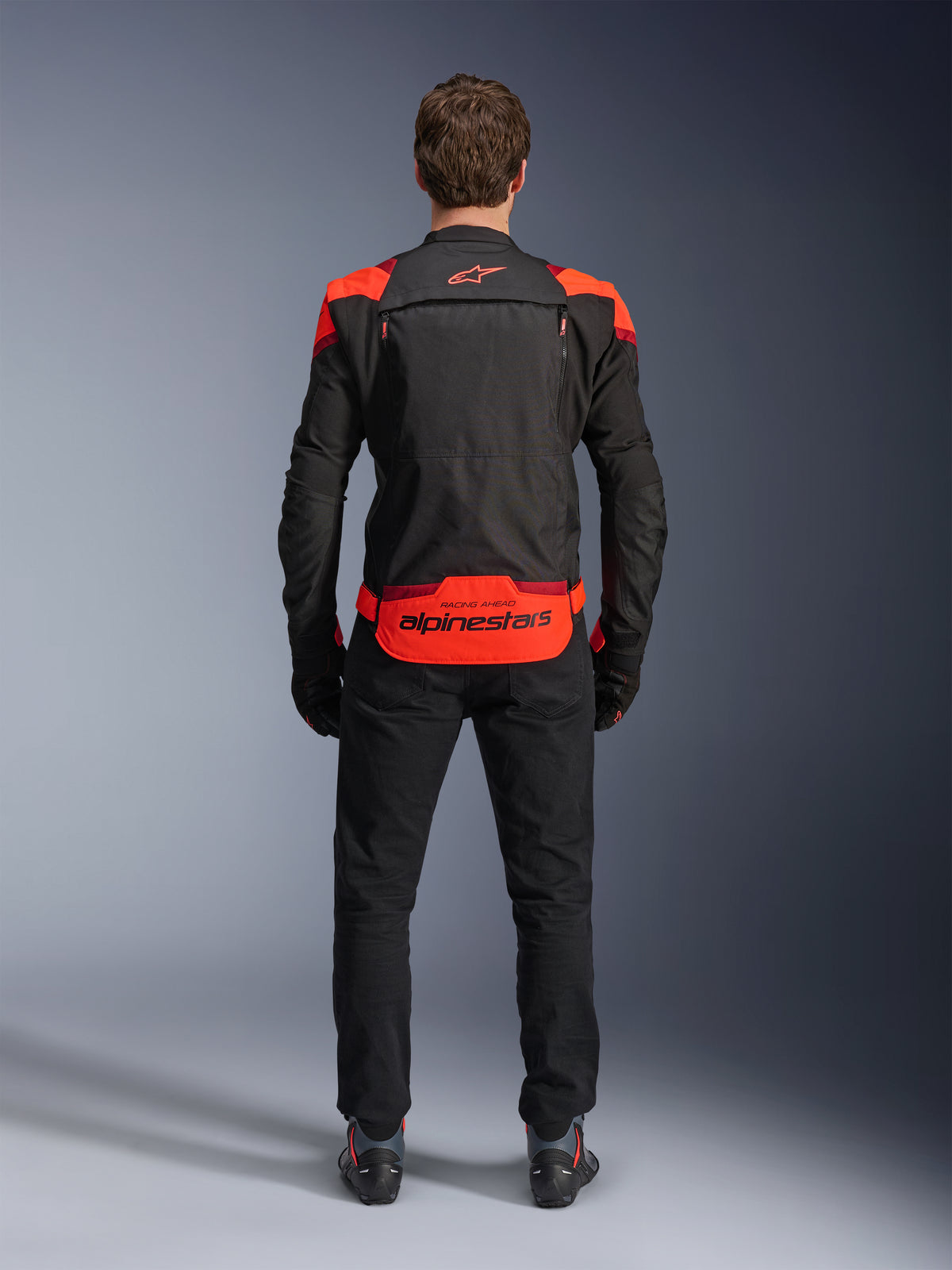 Alpinestars T-Stunt Air Jacke, Schwarz mit Red Fluo und dunkelroten Akzenten, Rückansicht am Model, kombiniert mit schwarzer Motorradhose und Stiefeln, markantes Alpinestars-Branding am unteren Rücken, modulares Design mit Mesh-Konstruktion für Belüftung